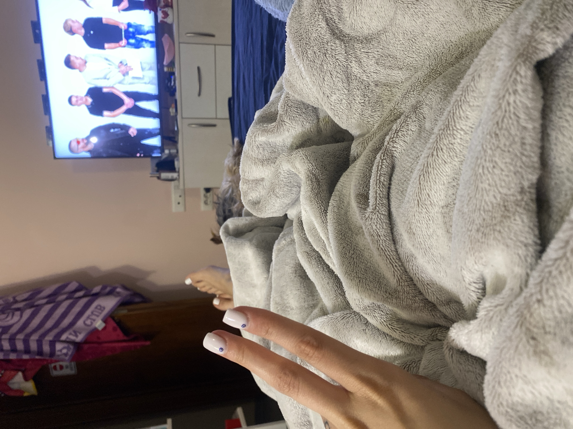 The image depicts an indoor setting. A person's hand is in the foreground with two fingers extended, the fingernails are painted white with small blue dots. A gray fluffy blanket dominates the right side of the frame. In the background is a television screen displaying four men. A white cabinet is visible to the right of the television. Below the hand is a dark wooden surface with some fabric or clothing with a striped pattern. To the right are visible toes with matching nail polish as the hands. The location is impossible to determine from the image alone. The city of Sofia, Bulgaria cannot be confirmed.