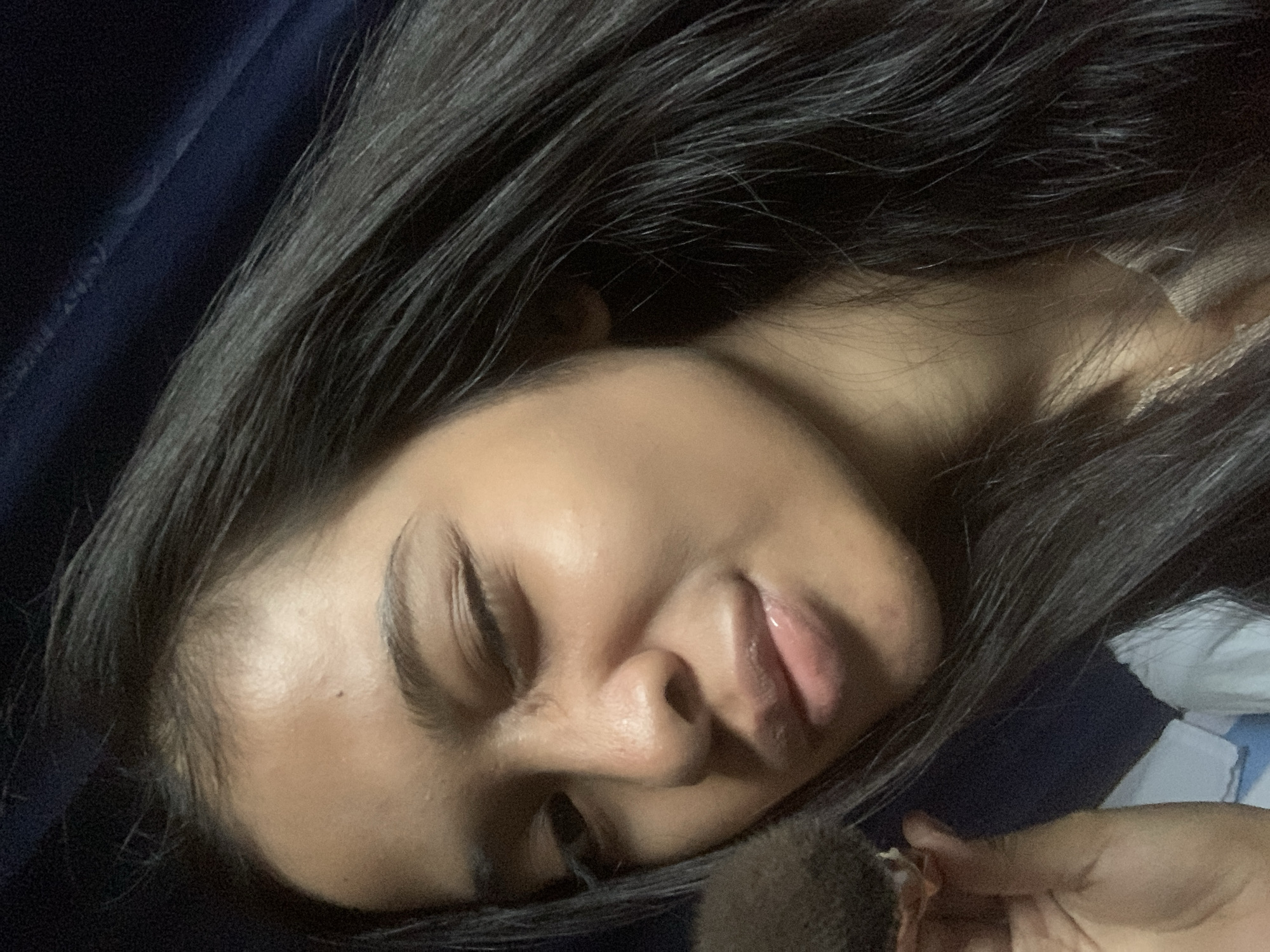 The image shows a close-up, overhead shot of a young woman with dark hair and medium-toned skin. She is winking with her left eye and looking towards the camera. Her lips are slightly parted and appear glossy. In the lower left corner, her left hand is visible, holding a fluffy, brown makeup brush near her cheek. She is wearing a light-colored garment that is partially visible. The background is dark, with a hint of a blue fabric to the left and top. There are no discernible landmarks or environmental features to indicate a specific location within Sungai Meriam, Indonesia.