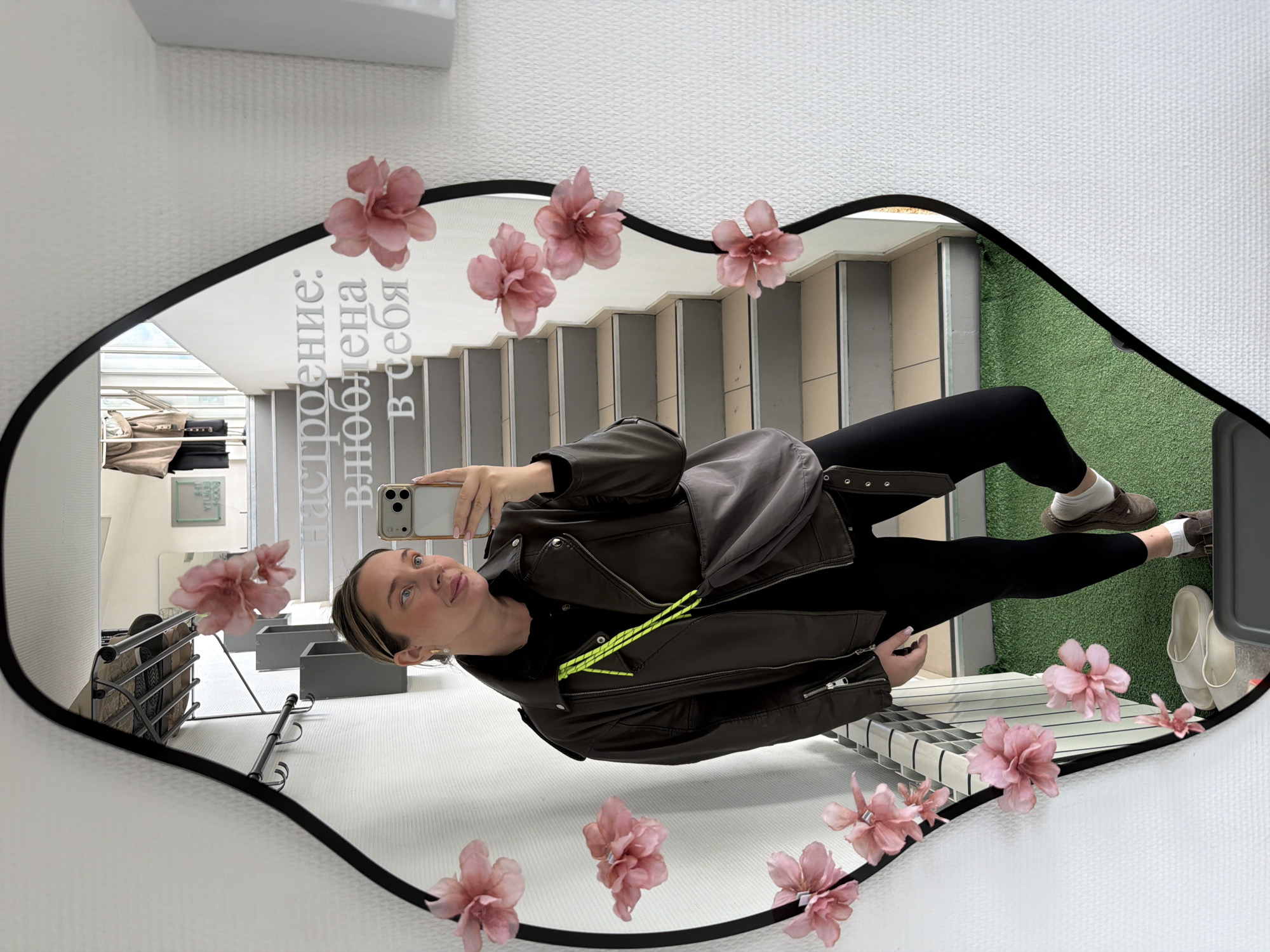 A woman is taking a selfie in a mirror in what appears to be a commercial space, potentially in the city of Chaikovsky, Russia, judging by the lettering in the reflection. The mirror, an irregular oval with a black frame, has several pink flower embellishments attached to it. The woman is wearing a black top, black leggings, brown slip-on shoes with white socks, and a brown leather jacket with a bright green decorative accent. Her phone is visible in her hand, positioned for the selfie. Reflected in the mirror are several elements including a staircase, an area with faux grass, shelving units, a white textured wall, and a window. The reflection also includes a slogan in Russian text.