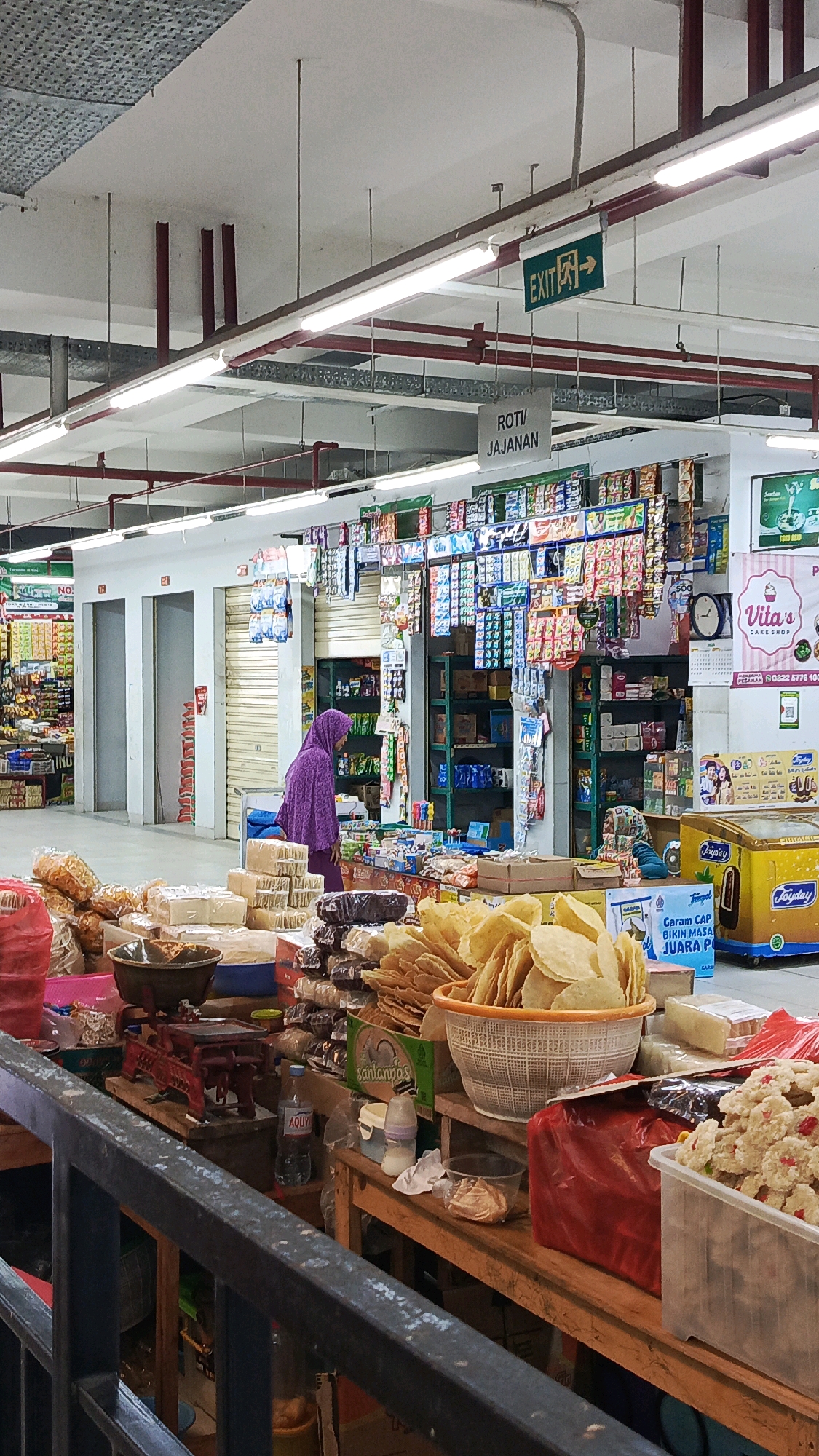 This image captures an indoor market scene, likely within the city of Ponorogo, Indonesia. In the foreground, a wooden market stall displays a variety of food items, including snacks like crackers or chips in a woven basket, packaged goods, and what appears to be a traditional red weighing scale. A woman wearing a purple hijab is visible in the mid-ground, browsing items at a stall filled with packaged snacks and drinks. Behind her, a row of closed shopfronts with rolling shutters suggests a typical market layout. Overhead, fluorescent lights illuminate the space, and an "EXIT" sign with directional arrows is prominently displayed on the ceiling. The environment appears to be a well-lit, bustling marketplace.