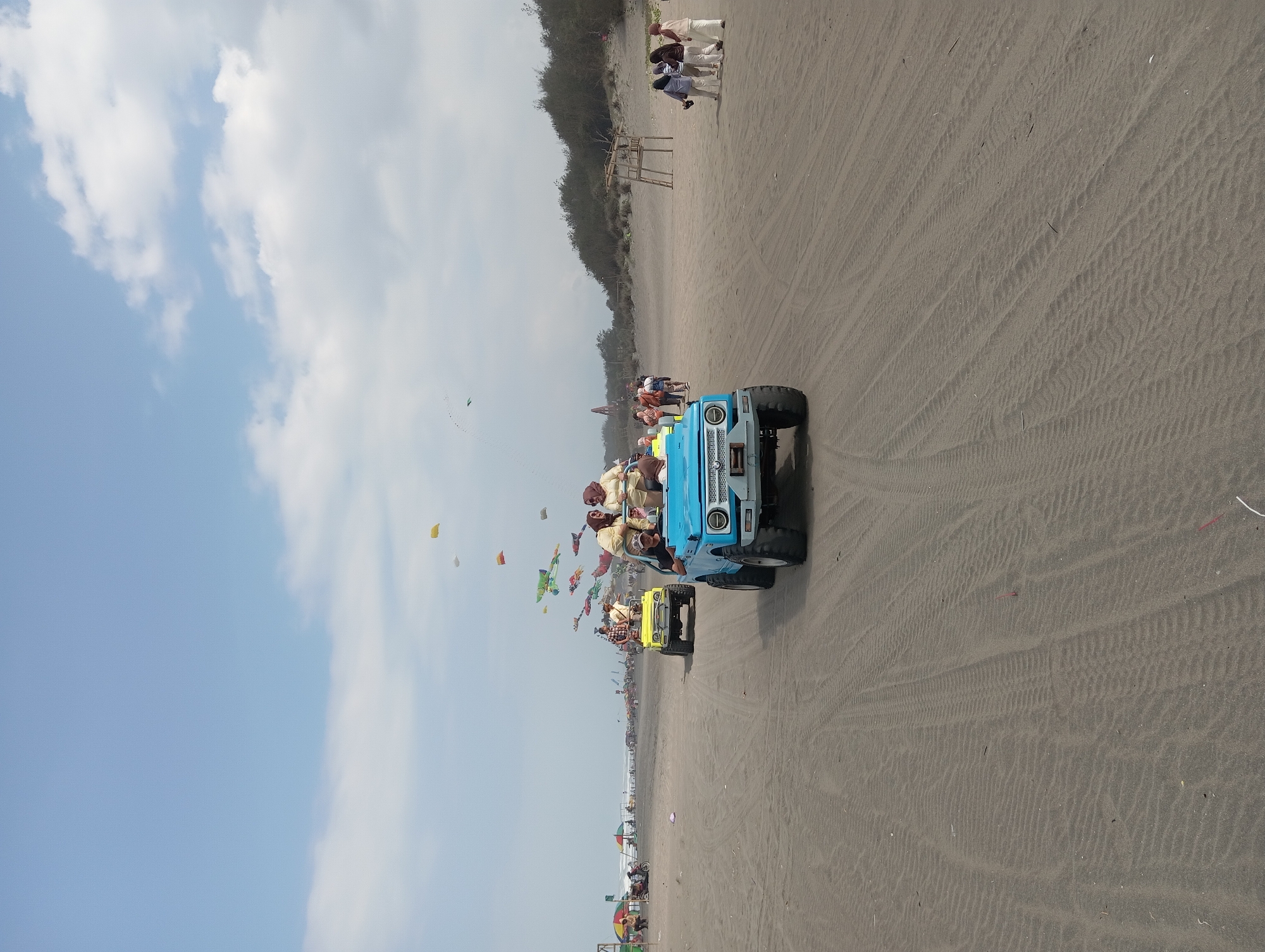The image depicts a sunny day on the sandy beach of Parangtritis, Indonesia. In the foreground, a blue, open-top jeep with several people riding in it is driving along the beach. Further back, a yellow jeep also with passengers is visible. The beach is populated with a scattering of people, some walking and some standing near the water's edge in the distance. The sky above is blue with scattered white clouds. Numerous colorful kites are flying in the sky, adding a festive element to the scene. The ground is covered in fine, rippled sand, with tire tracks from vehicles visible. To the right, a wooden structure on stilts stands on the sand dune, and a line of trees forms a backdrop. The overall impression is one of a leisure activity at a popular beach destination.