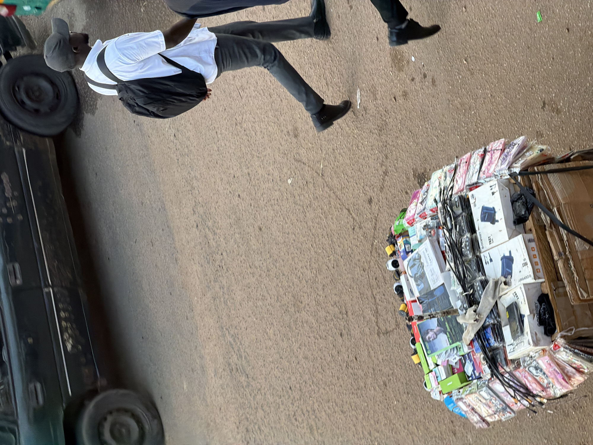 In Bukuru, Nigeria, a dark-colored car is parked on a dusty asphalt road, its paint weathered and scratched. In the foreground, a vendor's stall is piled high with various electronic accessories, including phone cases, chargers, and headphones. The items are a chaotic mix of colors and brands, displayed on what appears to be a wooden pallet. A man wearing a white polo shirt, dark trousers, and a black baseball cap walks past the stall, carrying a black backpack. His stance suggests he is moving with purpose. Another person, dressed in dark clothing, walks behind him, their feet shod in black shoes. The overall impression is a snapshot of daily life and commerce in a Nigerian urban setting.