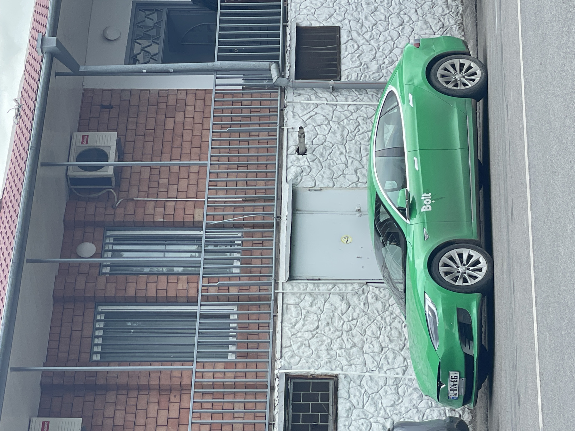 A green Bolt-branded Tesla car is parked on the street next to a multi-story building. The building has brick and white stone sections, metal grates over the windows, and an air conditioning unit. The license plate of the Tesla is visible, reading "MJ-094-GG." Based on the car and architectural styles, this image could be taken in Tbilisi, Georgia.