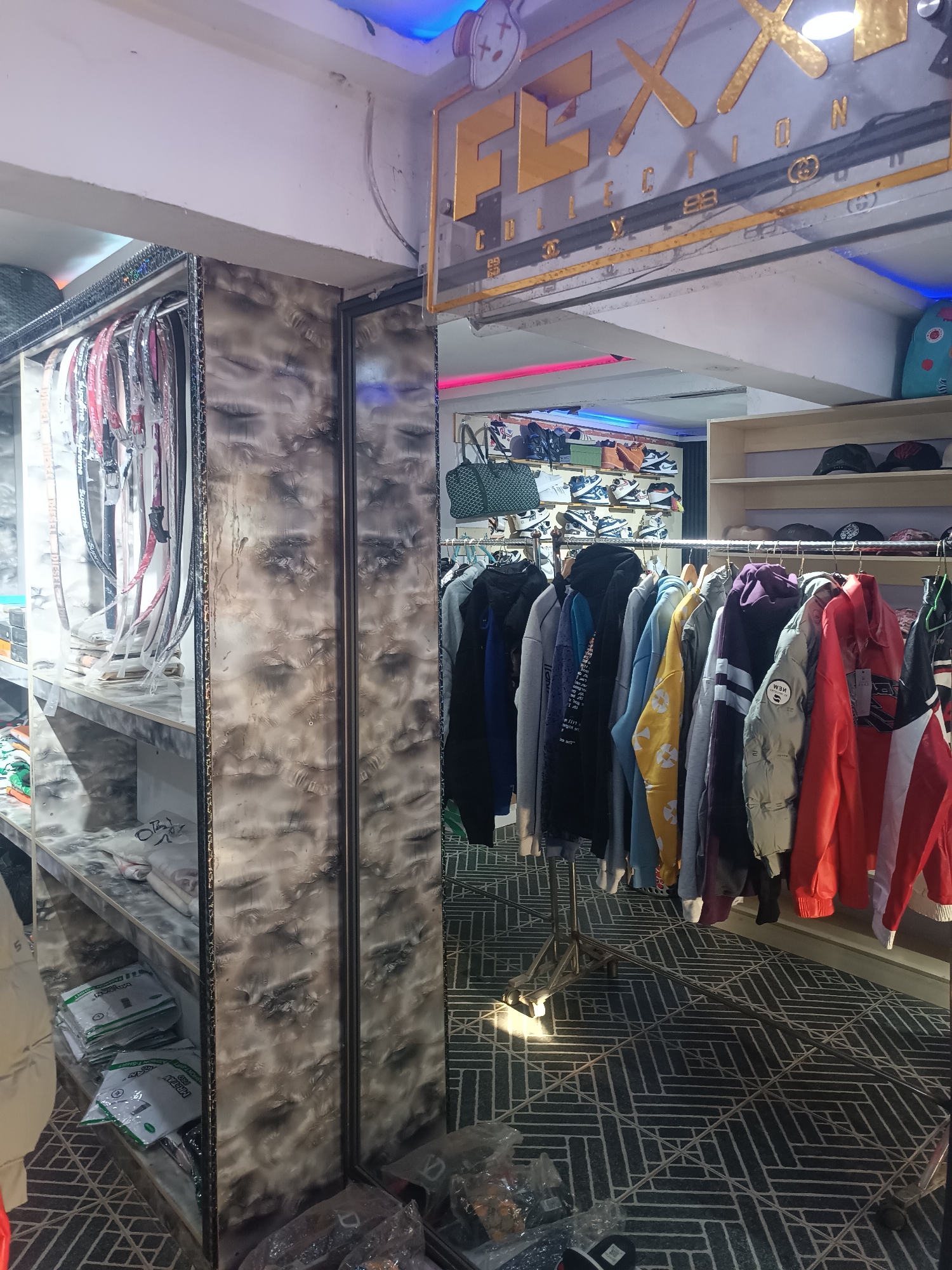 The image shows the interior of a retail store, likely a clothing boutique in Bukuru, Nigeria. The store's entrance is marked by a sign with the words "FLXX COLLECTION," written in gold lettering. To the left, a mirrored display case houses belts and packaged items on shelves. Racks of clothing, including hoodies and jackets in various colors, are displayed on the right. In the background, there is a display of shoes and caps. The floor has a geometric pattern. There are no people visible in the image.