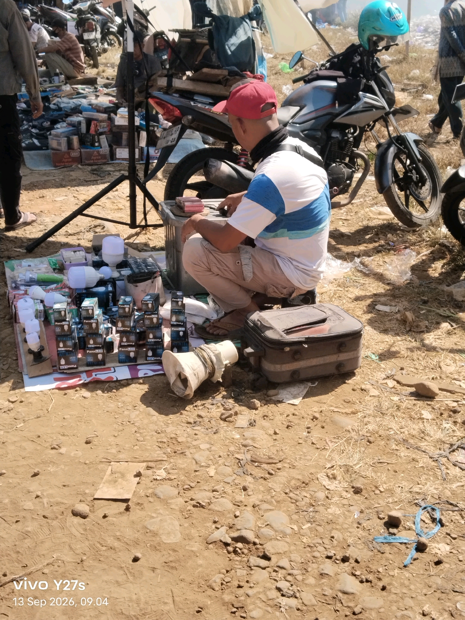 The image depicts an outdoor marketplace, likely in Wonosido, Indonesia, with a focus on a vendor selling electrical components. A man wearing a red cap and a blue and white striped shirt is kneeling beside a display of LED light bulbs and their packaging. The bulbs are arranged on a white tarp spread on the ground, alongside various boxes, possibly for sale. In the background, motorcycles are parked, and other individuals are visible, suggesting a busy market environment. The ground is dirt and gravel, consistent with an outdoor setting. A black case with a red accent sits in front of the kneeling man. The overall scene conveys a sense of commerce and everyday life in a local Indonesian market.