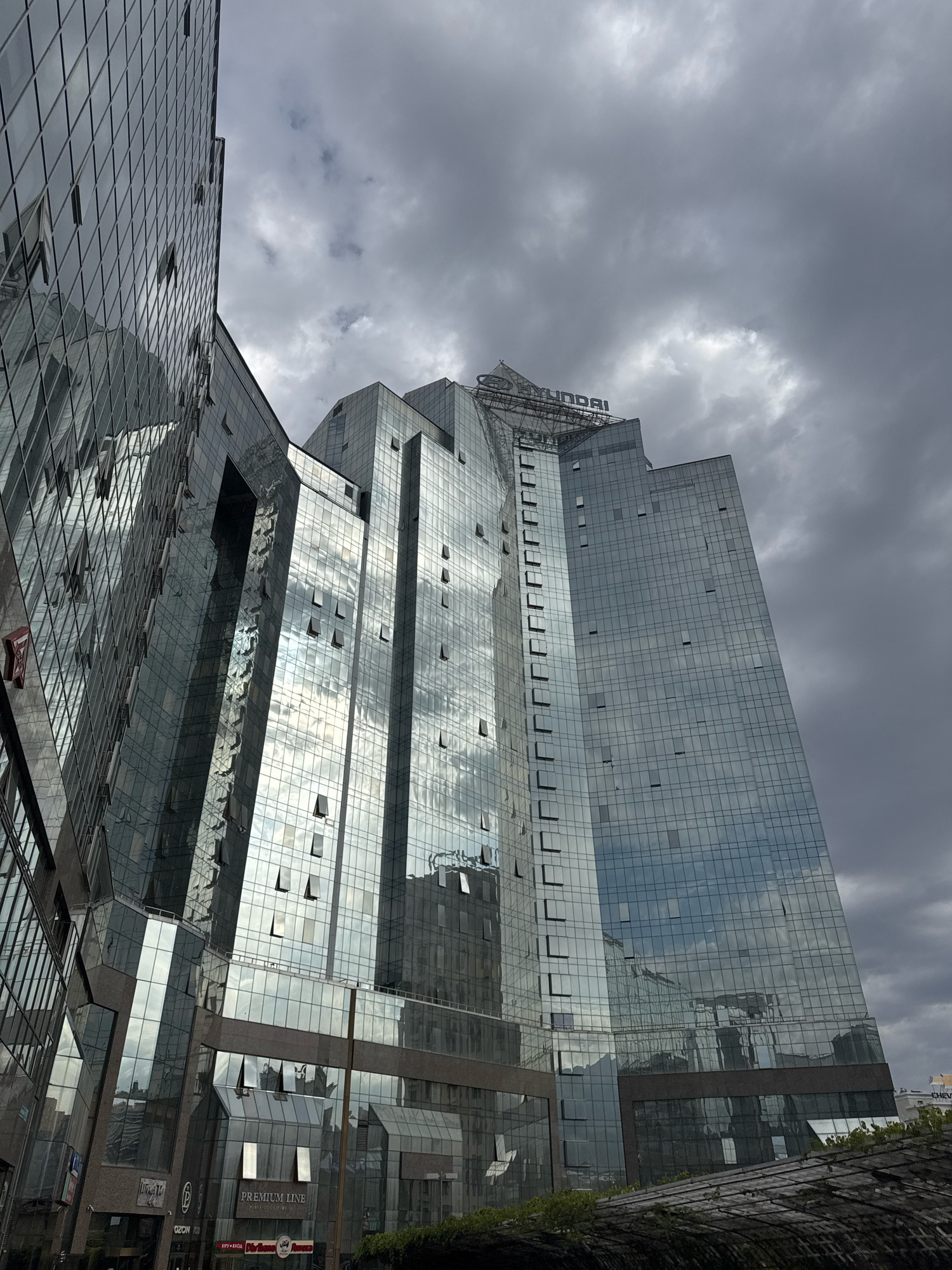 The image captures the exterior of the Hyundai Central Asia building in Almaty, Kazakhstan. The skyscraper, with its modern glass facade, dominates the frame, reflecting the overcast sky and surrounding buildings. Some of the windows on the skyscraper are open. The building is situated in a dense urban environment, with other buildings visible in the background. The overall impression is one of a modern, functional office building against a backdrop of a cloudy day in a city.