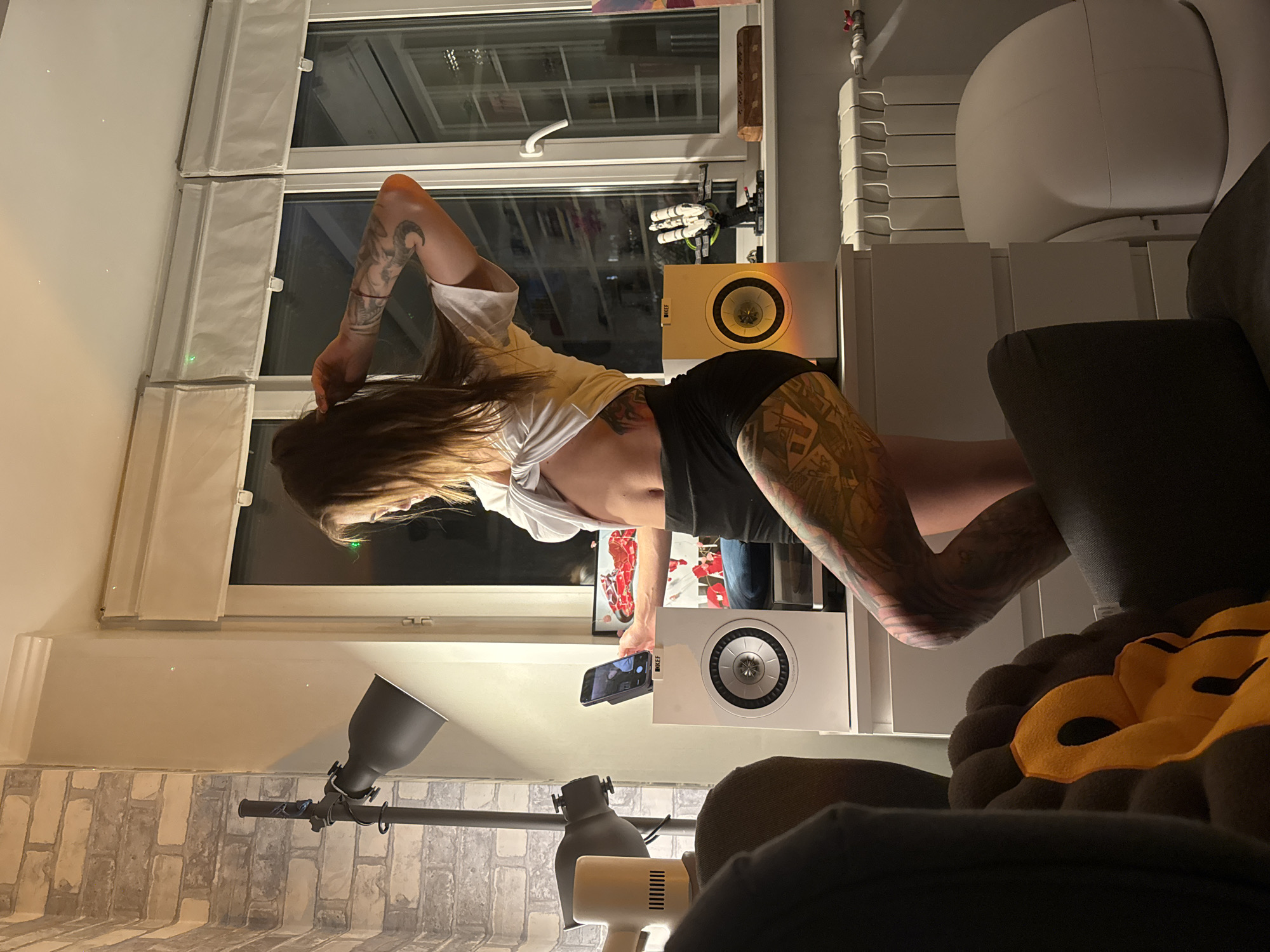 A young woman with extensive tattoos on her arms and legs is standing in an apartment. She is wearing a white t-shirt, tied at the front to reveal her midriff, and black shorts. Her right hand is raised, touching her hair, as she looks down at her phone, which is placed on a white speaker. Next to this speaker is another speaker with a yellow grille. In the background, there are large windows with white blinds, a brick-patterned wall, and a floor lamp. The room appears to be a living room or bedroom, with furniture including a dresser, a radiator, and part of a sofa in the foreground. The lighting is dim, suggesting it is evening or night. The provided coordinates indicate this location is in Moscow, Russia.