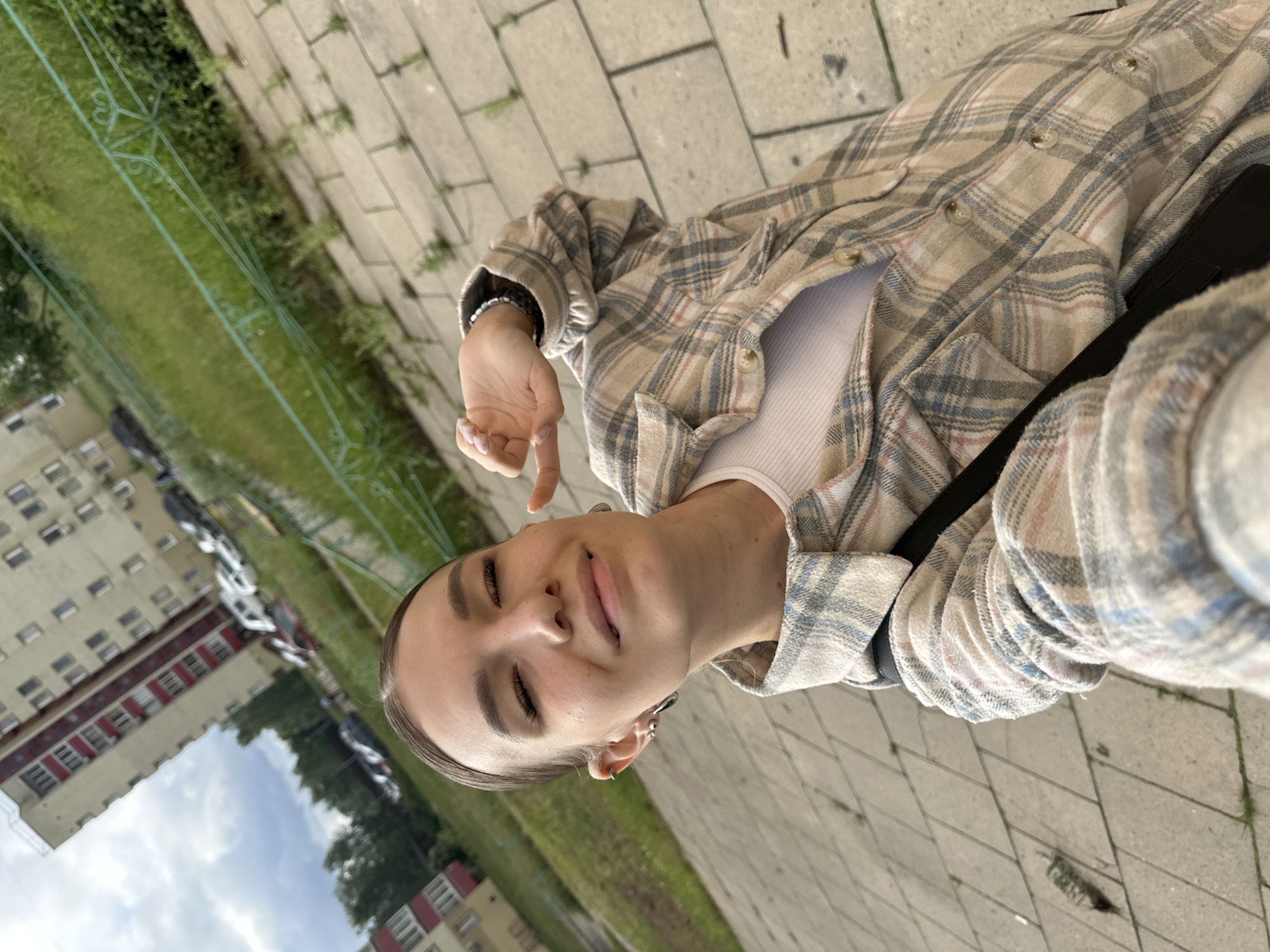 A young woman with dark hair pulled back into a bun and closed eyes is taking a selfie outdoors. She is wearing a white tank top and a plaid button-up shirt in beige, blue, and pink. Her left hand is raised and positioned near her face, obscuring part of it. She is wearing earrings and a bracelet. The background shows a paved walkway with some grass growing between the stones, a green metal fence, a grassy area with trees and parked cars, and buildings in the distance. The scene appears to be in a residential area within the city of Chaikovsky, Russia.
