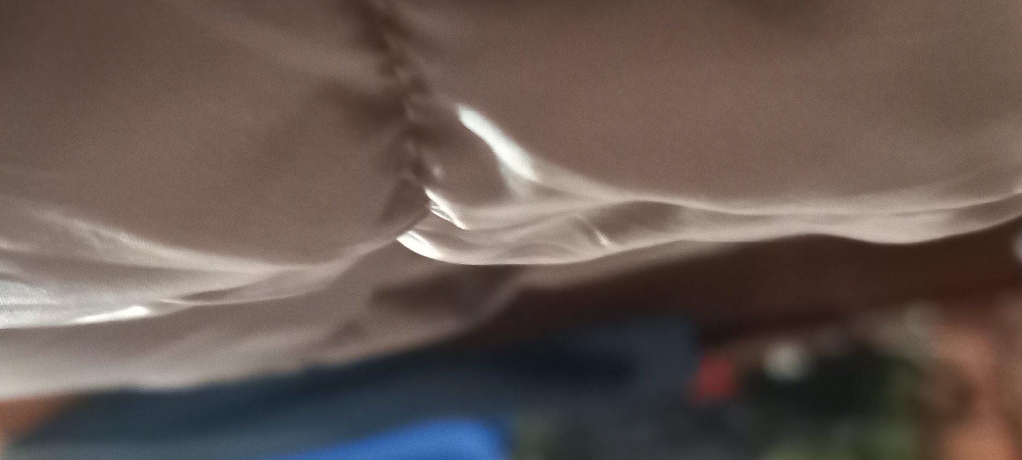 This abstract image presents a close-up view of a light-colored, possibly beige or cream, fabric. The material is characterized by numerous soft folds, wrinkles, and deep shadows, creating a sense of depth and texture. Bright, elongated highlights streak across the fabric, indicating a strong, unseen light source, likely daylight entering an indoor space.
Towards the upper-left and center of the frame, a distinct seam or a gathered, ruched section of the fabric is visible, contributing to the fabric's complex topography. The bottom portion of the photograph features a heavily blurred and out-of-focus background, showing indistinct shapes and colors. These blurred elements include shades of dark blue, brown, and subtle hints of red and green, suggesting the presence of furniture or other objects in a room.
There are no people or specific activities taking place in the image. The scene is static, focusing purely on the interplay of light and shadow on the textile. No visible text is present. The general setting appears to be an indoor environment, given the nature of the fabric and the blurred background elements. The image itself does not contain specific cues to its location in Kryvyi Rih, Ukraine, beyond the provided context.