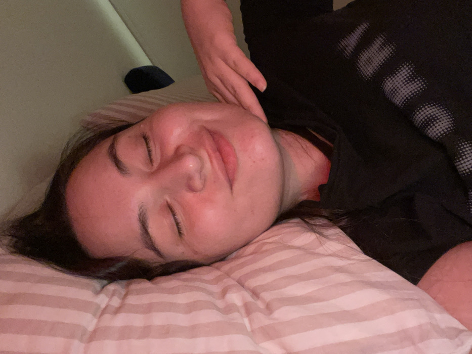 A woman with closed eyes rests on a striped pillow. Her skin appears slightly flushed, and she's wearing a black shirt. One hand is gently placed near her face. The background includes a wall. The lighting is soft and subdued. The image focuses primarily on the woman's peaceful expression.