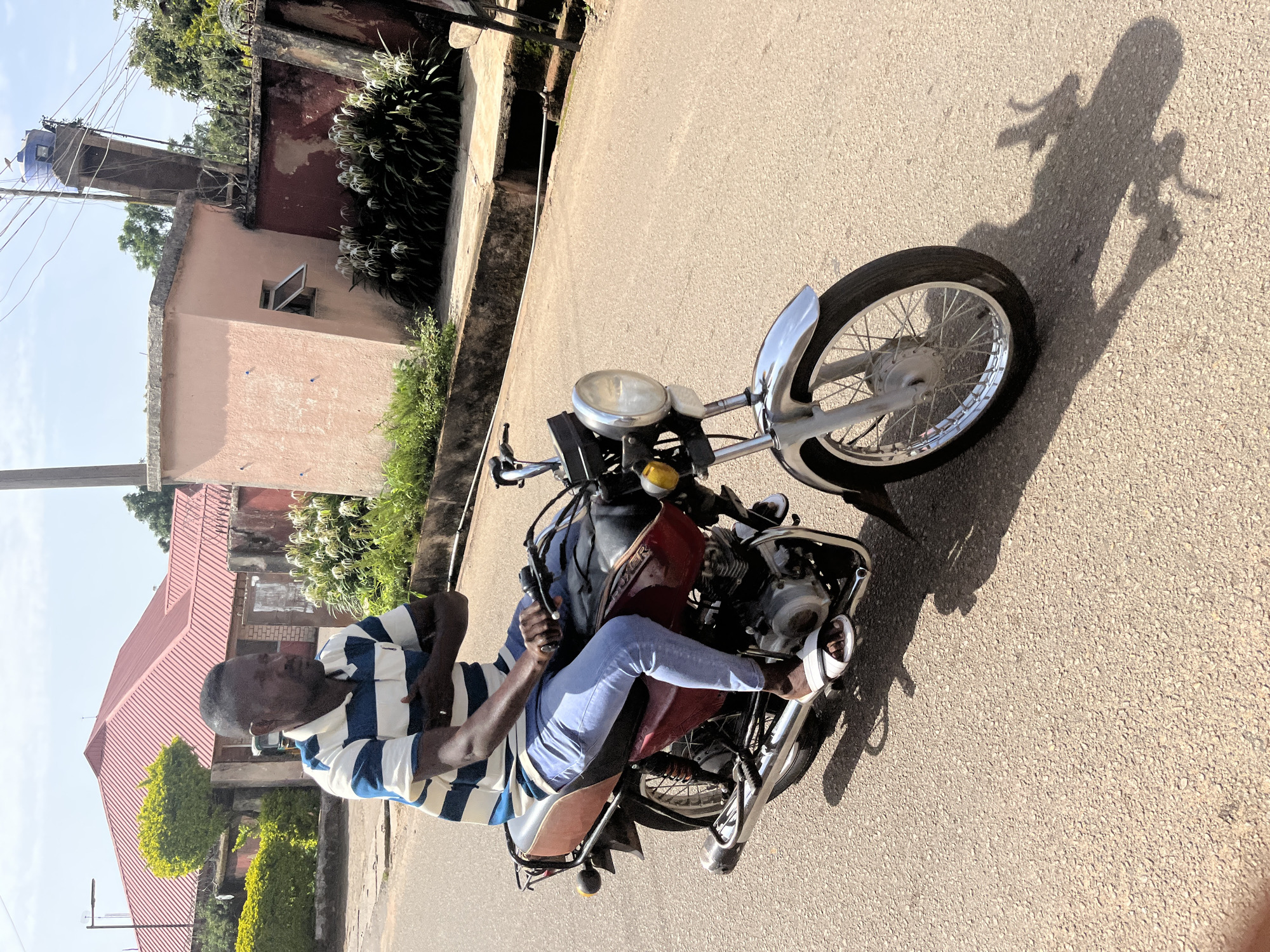 A man is riding a motorcycle on an asphalt road in Jos, Nigeria. He is wearing a blue and white striped t-shirt and light blue jeans. The man is looking to his right and has his arms crossed. The motorcycle is dark red with a brown seat and chrome accents. In the background, there are houses, trees, and a blue water tank on a tower. The sky is clear and the sun is shining.