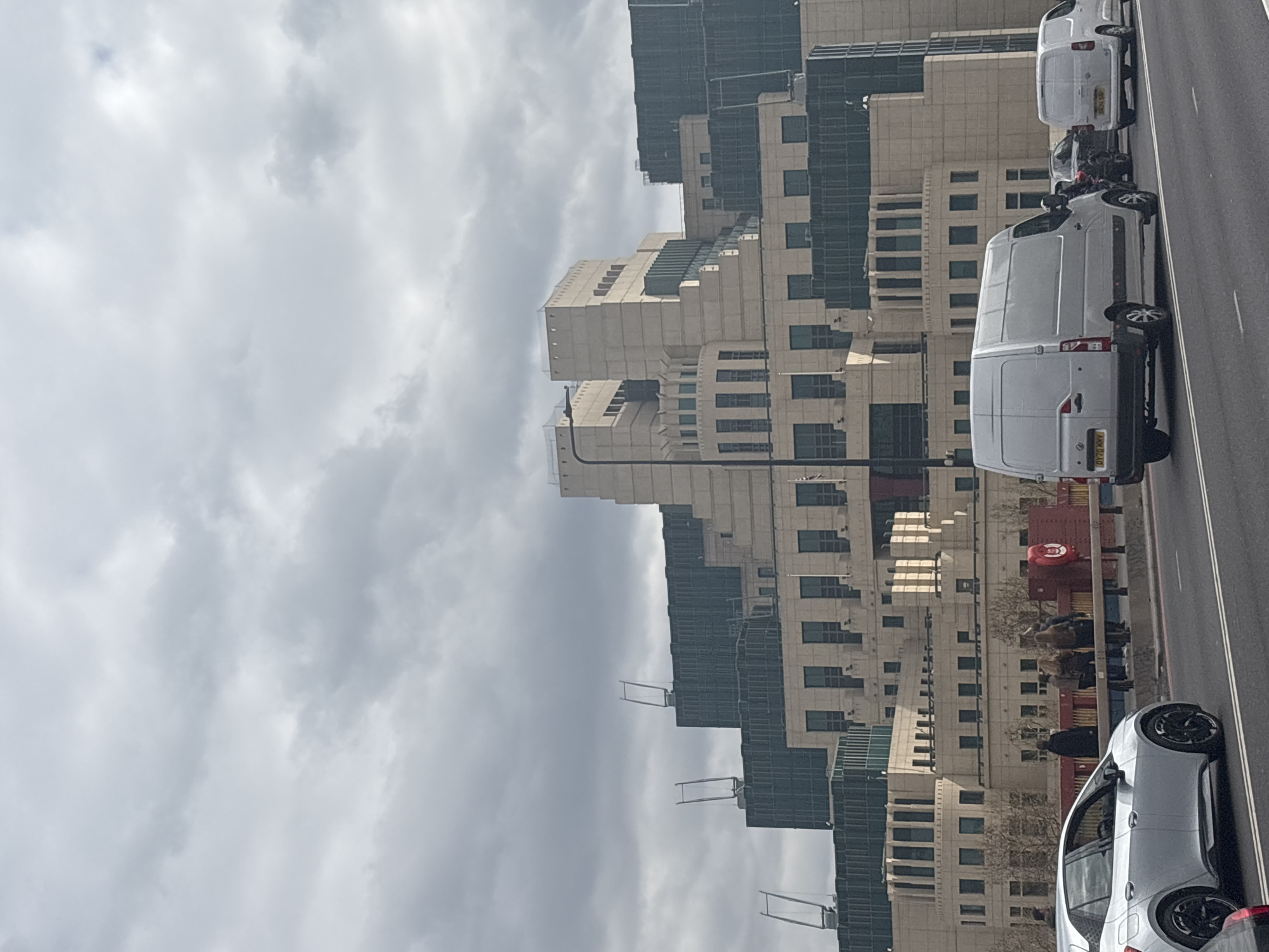The image depicts a street scene in London, United Kingdom, likely near Vauxhall Bridge. The SIS Building, also known as the MI6 Building, is visible in the background. A road runs alongside the building, with vehicles including white vans and a silver car parked or driving on the road. Pedestrians are present on the pavement adjacent to the road. The sky is overcast with heavy cloud cover.