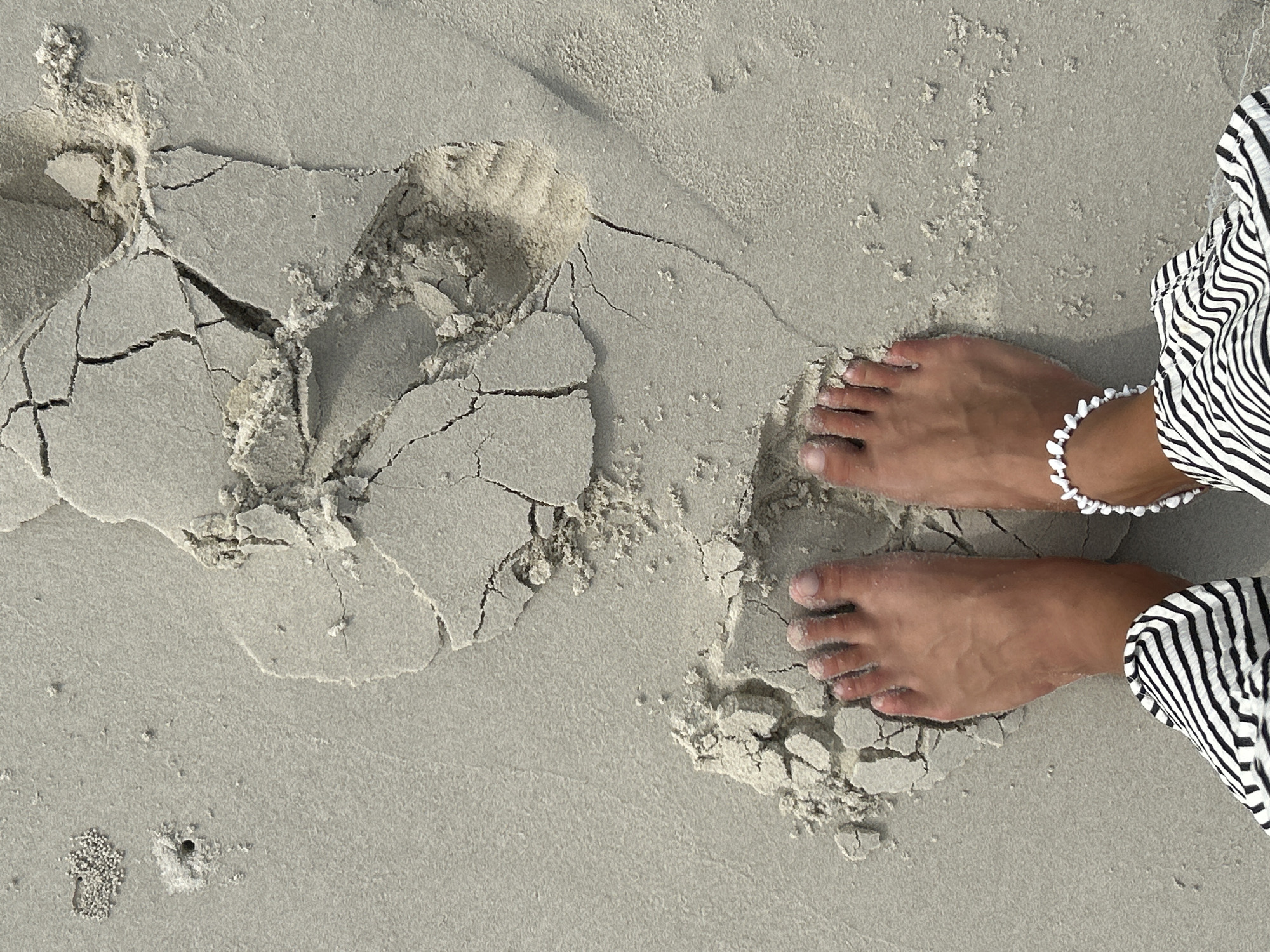 A pair of bare feet, adorned with a white beaded anklet on the right ankle, are standing on a light-colored sandy beach. The feet are partially covered in fine sand. The sand is textured and shows several cracks and broken fragments, suggesting it may have been recently wet and dried. In the upper left quadrant of the image, there is a small hole in the sand surrounded by a cluster of granular material, possibly indicating the presence of a small marine creature. The background is a smooth expanse of sand. The lower portion of the image shows the black and white striped fabric of clothing. The scene is set in Ko Yao, Thailand, on a beach.
