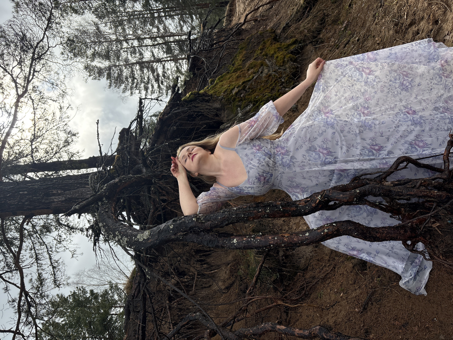 A young woman with light-colored hair is depicted lying serenely among large, dark, gnarled tree roots in an outdoor setting. She is wearing a long, flowing dress adorned with a delicate floral pattern in shades of lavender, purple, and white, featuring sheer sleeves. Her eyes are closed, and her face is angled slightly upwards, conveying a peaceful or contemplative expression. Her left hand rests near her temple, while her right hand gently touches the fabric of her dress.
The scene takes place in a natural environment, likely a forest or a bank with exposed earth and tangled roots, which are prominent in the foreground and around the woman. Tall trees, possibly pines or birches, stand in the background against an overcast or cloudy sky, suggesting a muted, diffused light typical of daytime. The ground beneath the roots appears to be sandy or earthy. The overall atmosphere is tranquil and somewhat ethereal, highlighting a connection between the woman and nature. No text is visible in the image. The location is Chaikovsky, Russia.