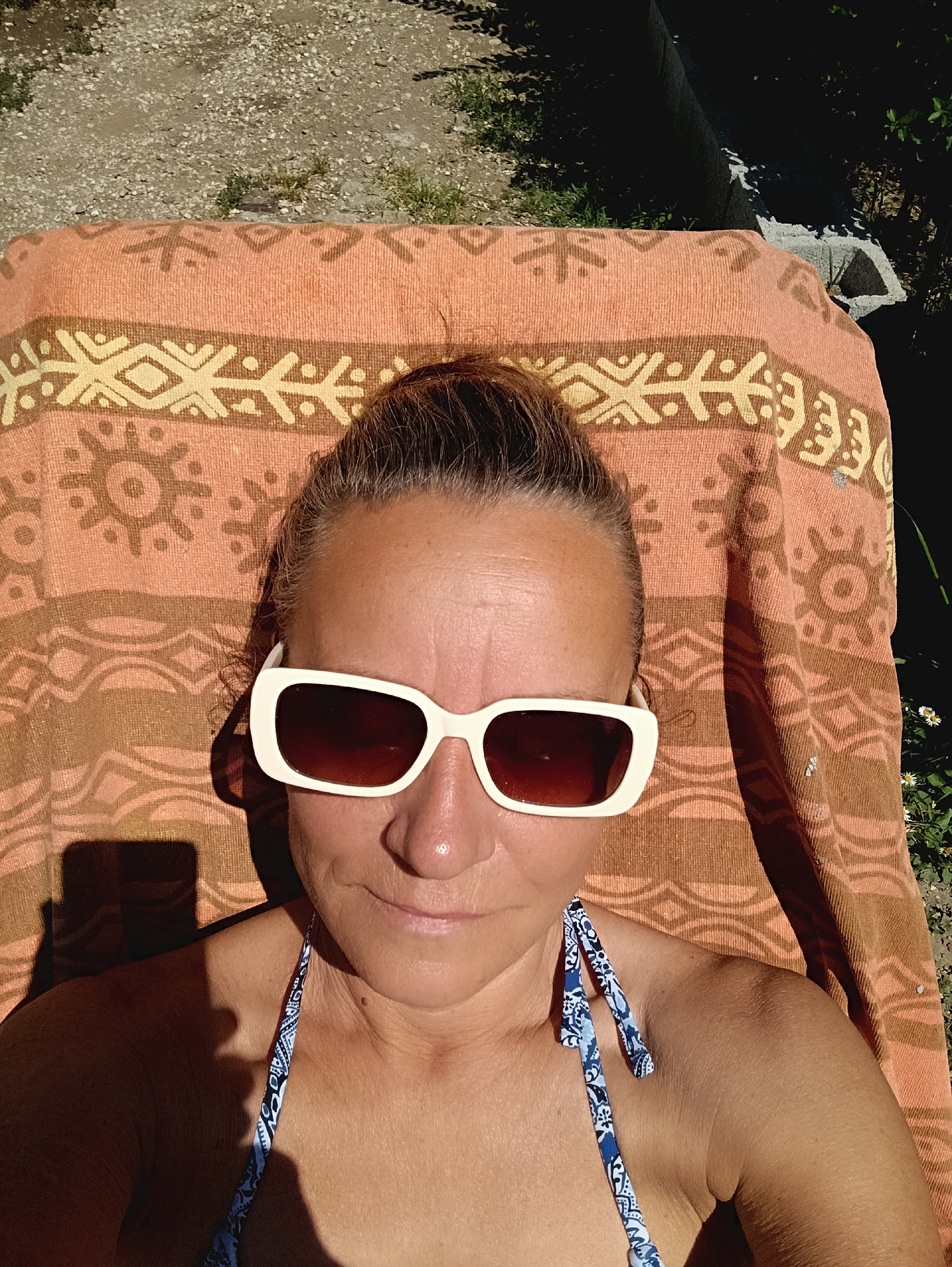 A woman wearing sunglasses and a swimsuit lies on a patterned orange and brown towel. The woman's hair is pulled back in a bun. The location is outdoors, seemingly in a garden or yard, possibly in the city of Viadana, Italy. A concrete block is visible in the background, with greenery and flowers. Strong sunlight casts shadows on the woman's face and body.