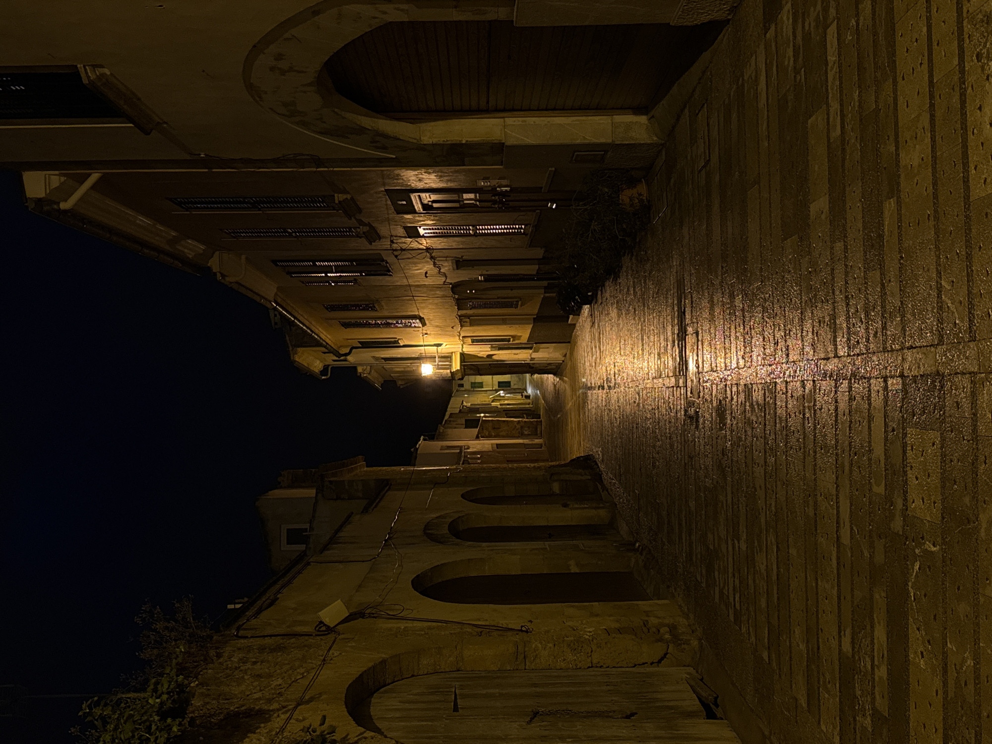 The image depicts a narrow street at night in what appears to be Alcúdia, Spain. The street is flanked by tall, old buildings with windows and shutters. The walls are made of stone and illuminated by dim, warm streetlights, and some windows show signs of light. The street surface appears wet. In the distance, the street ascends a slight incline. Arches are visible at the bottom of the frame, close to the street, and one can spot some wiring hanging at the right. There is no presence of people or animals. The sky above is a dark, clear night.