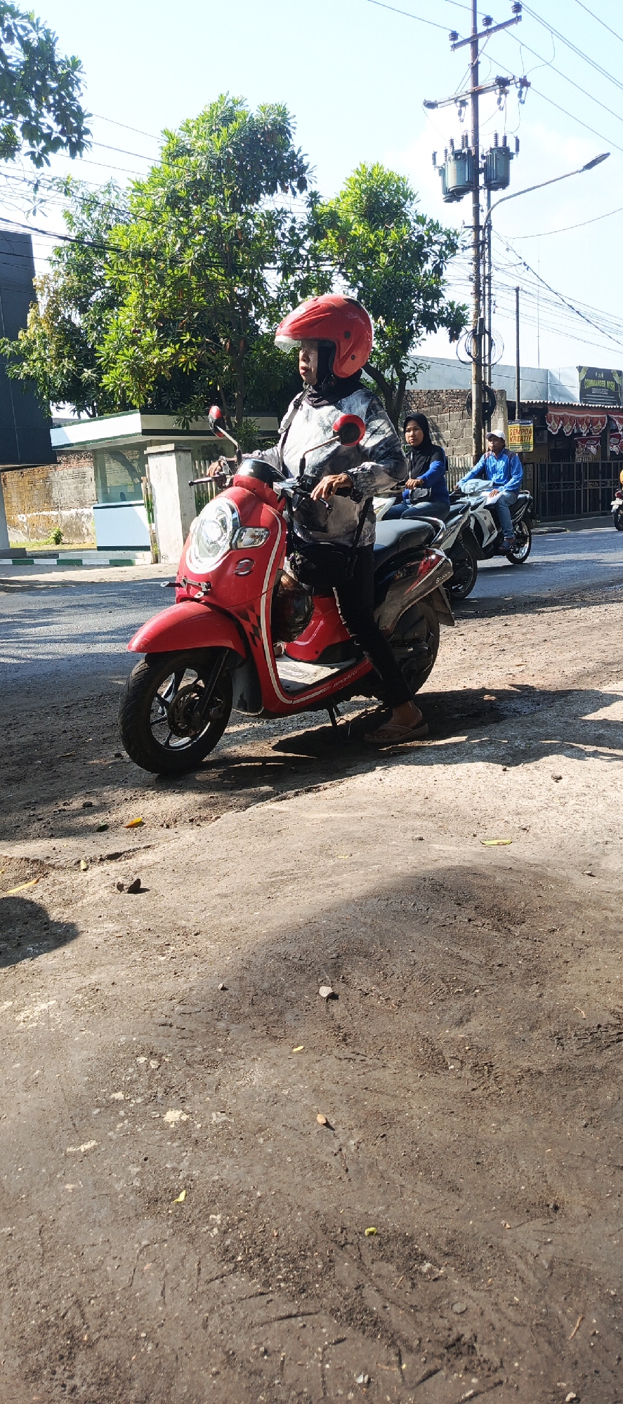 An outdoor, medium shot captures a red scooter with a rider in a red helmet. The rider, wearing a patterned jacket and black pants, is seated on the scooter, holding the handlebars. The scooter is positioned on a dirt and asphalt surface, suggesting a roadside or parking area. In the background, a utility pole with transformers and wires stands tall. Trees with green foliage flank the scene, and buildings with signs are visible further in the distance. Two other individuals are on motorcycles behind the main rider, one wearing a blue jacket and the other a black hijab. The lighting indicates a sunny day, casting shadows on the ground. The scene appears to be a street in Krian, Indonesia, with the utility pole and style of buildings characteristic of the region.