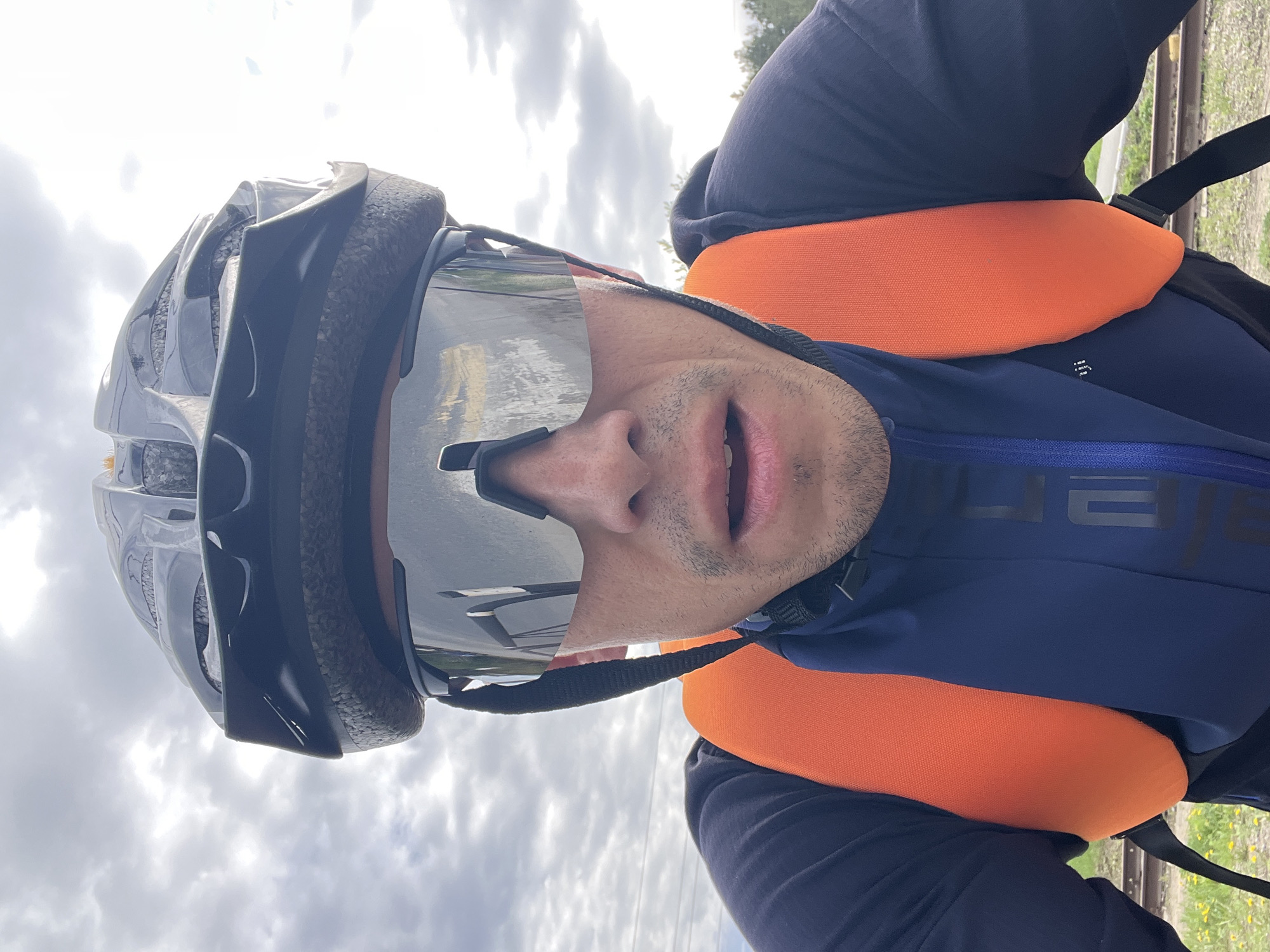 A man wearing a black helmet, reflective sunglasses, and a blue jacket with orange straps is positioned against a cloudy sky. The straps seem to be part of a life vest or similar buoyant device. His mouth is slightly open. The location appears to be near railroad tracks. The location is the city of Chaikovsky, Russia, based on information requested in the prompt.