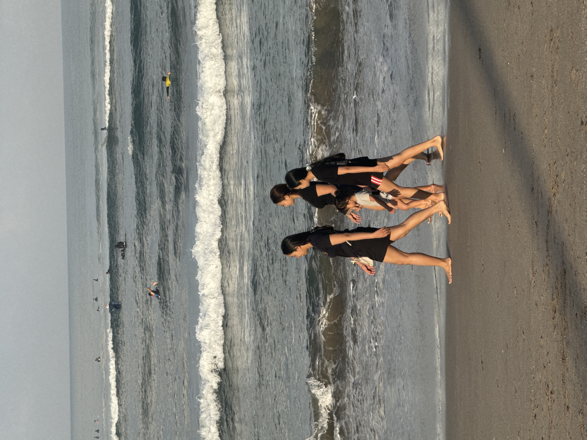 At a beach in North Kuta, Indonesia, four people are walking along the shoreline. Two adult women and two young children are walking from right to left. The adults are wearing black shirts and shorts, while the children are in lighter-colored clothing. In the background, the ocean has moderate waves, with several surfers visible in the distance. The sky is overcast, and the overall lighting suggests it is late afternoon or early evening. The sand is wet and dark near the water's edge, gradually becoming lighter and drier further up the beach.