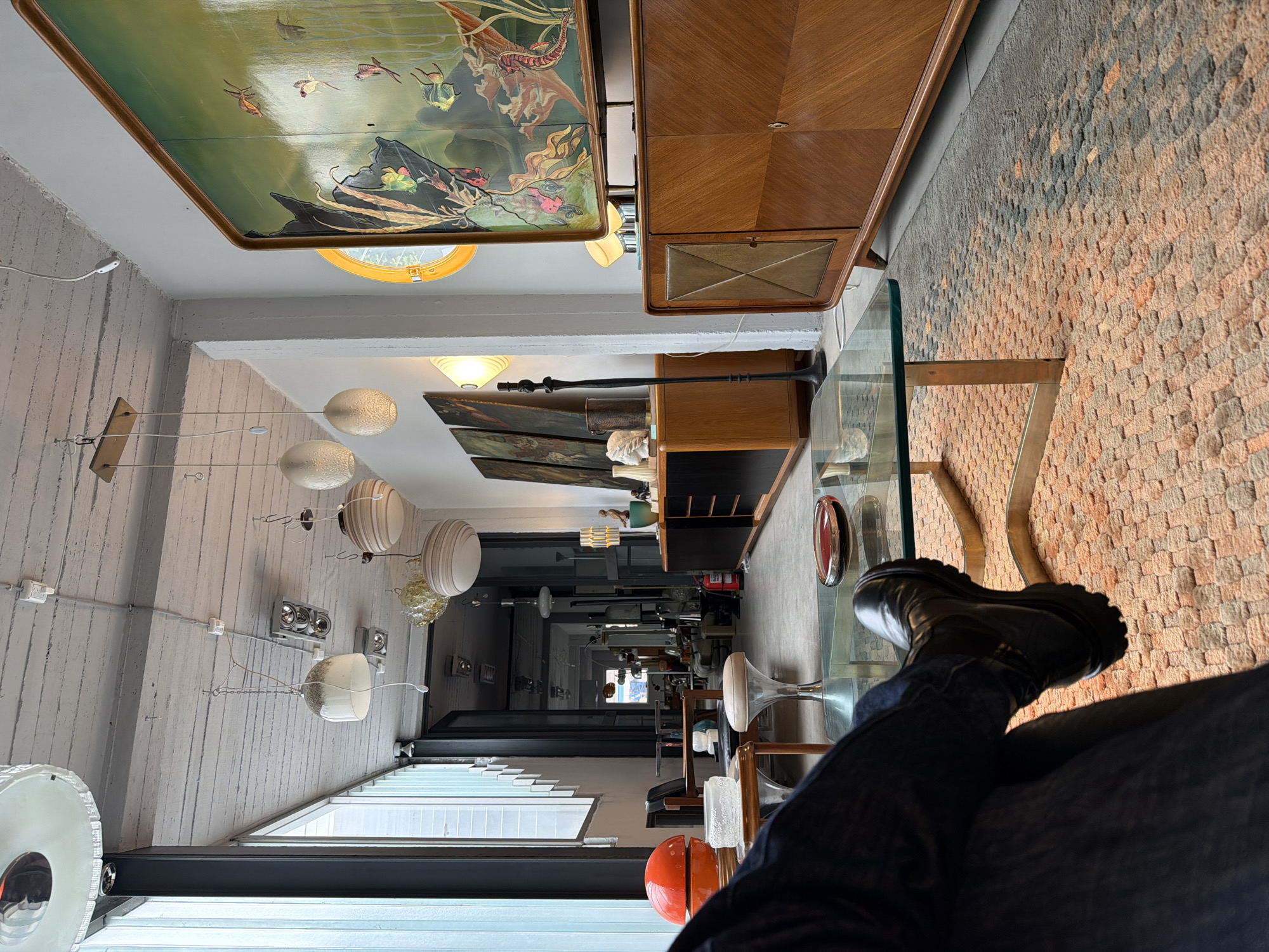 The image captures an indoor scene, likely a showroom or gallery, viewed from a seated perspective. The foreground features a person's legs, clad in dark jeans and sturdy black boots, resting on or near a glass-top table.
The room exhibits an eclectic mix of furniture, lighting, and art. On the left, a textured white wall with vertical plank-like patterns is adorned with multiple hanging light fixtures. These include frosted spherical lights and cylindrical lights with horizontal stripes, connected by exposed wiring and conduits. Further along this wall, a window with horizontal blinds allows natural light into the space. In the lower-left corner, a part of a red/orange spherical object is visible.
Above, or on an adjacent ceiling/wall, hangs a large rectangular artwork or decorative panel featuring a vibrant underwater scene with fish, seahorses, and aquatic plants. This piece is framed in wood and reflects light. A round yellow window or light fixture is visible near this artwork.
To the right, a distinctive wooden cabinet with geometric wood grain patterns and a central metallic inset door stands prominently. Further into the room, another wooden cabinet is visible, topped with a tall, slender black decorative object, possibly a lamp or sculpture. The floor appears to be concrete.
The far right wall features a rough, textured brick surface with varying shades of red, orange, and gray, suggesting an exposed or decorative brick treatment. Another glass-top table is partially visible in front of this wall.
The space extends into a long corridor or open area, revealing more furniture pieces, light fixtures, and art, creating a sense of depth. The overall impression is a curated space, possibly a vintage furniture store or a designer's studio, in Buenos Aires, Argentina, brightly lit, likely during the daytime. No explicit text is visible in the image.