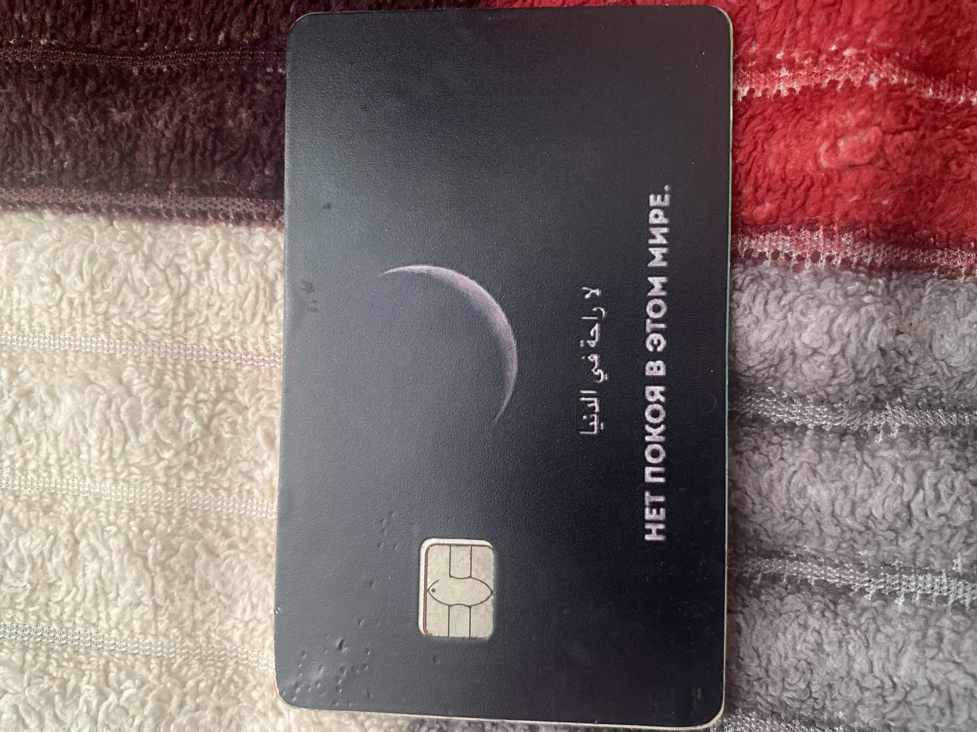 The image shows a black credit card with a chip on a textured surface. The card features a crescent moon graphic and text in Arabic and Russian. The Arabic text reads "لا راحة في الدنيا" (La raha fi al-dunya), which translates to "No rest in this world." The Russian text below it says "НЕТ ПОКОЯ В ЭТОМ МИРЕ" (NET POKOYA V ETOOM MIRE), also meaning "No rest in this world." The credit card is lying on a soft, fuzzy material that appears to be a blanket or rug. The background consists of a textured fabric in shades of cream, burgundy, and red. There are no people or specific environmental elements identifiable to pinpoint a location in Kizilyurt, Russia, or any other city. The scene is a close-up shot of the credit card against the textured background.
