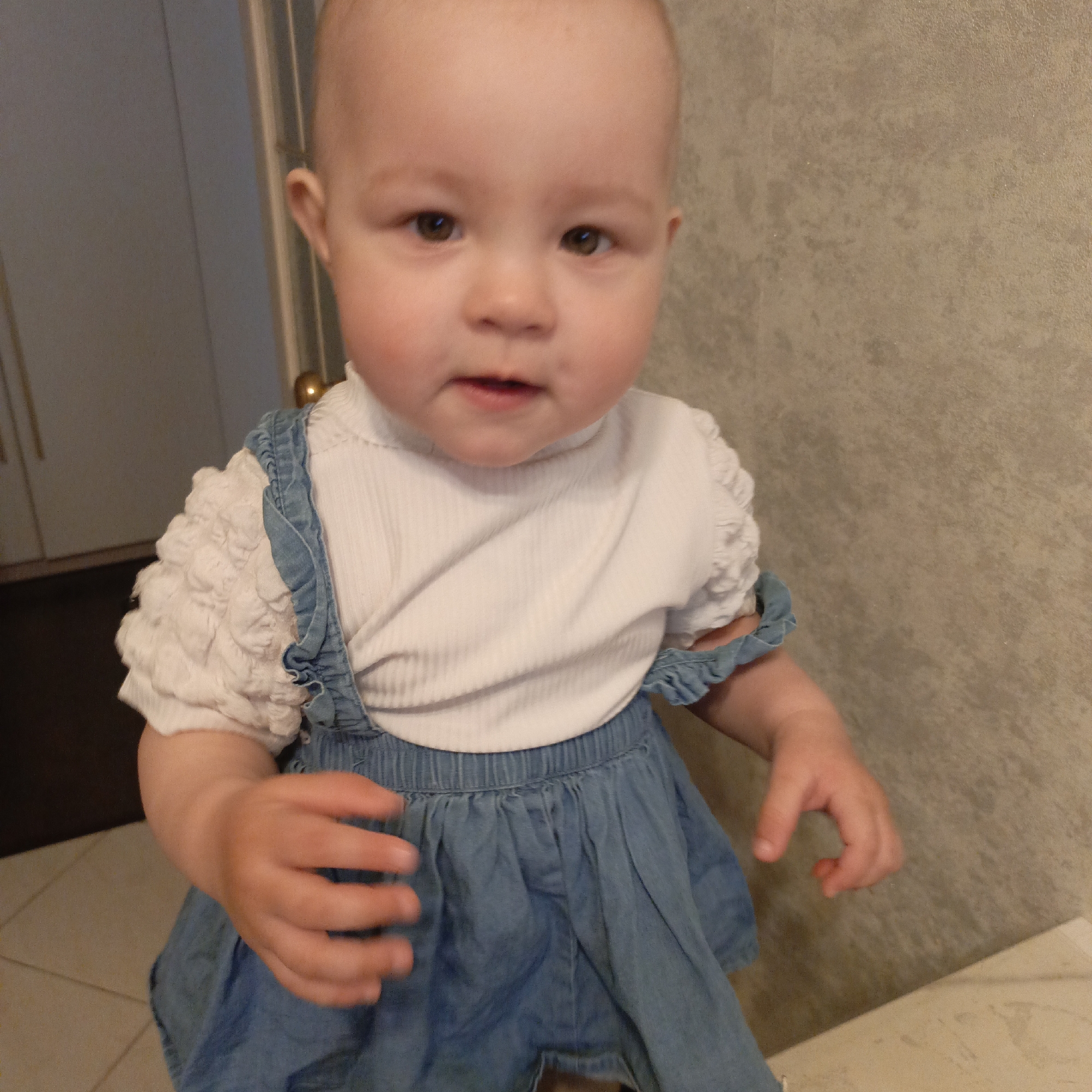 A young baby or toddler is pictured standing indoors, looking directly at the camera with wide, attentive dark brown eyes and a neutral, slightly curious expression. The child has fair skin and a bald head. They are dressed in a white, ribbed turtleneck top with short, puffy, textured sleeves, layered under a light blue denim-style romper or overall dress with shoulder straps and a gathered skirt.
The background on the left reveals a dark brown or grey door with a gold doorknob, next to a tiled floor. To the right and behind the child is a textured wall in a muted beige or brown tone. A light-colored surface, possibly a counter or furniture, is visible in the lower right foreground. The baby's left hand is raised with fingers slightly spread, suggesting movement or interaction, while the right hand is curled at their side. The scene is brightly lit, consistent with an indoor setting during the day or under artificial light. No text is visible in the image.