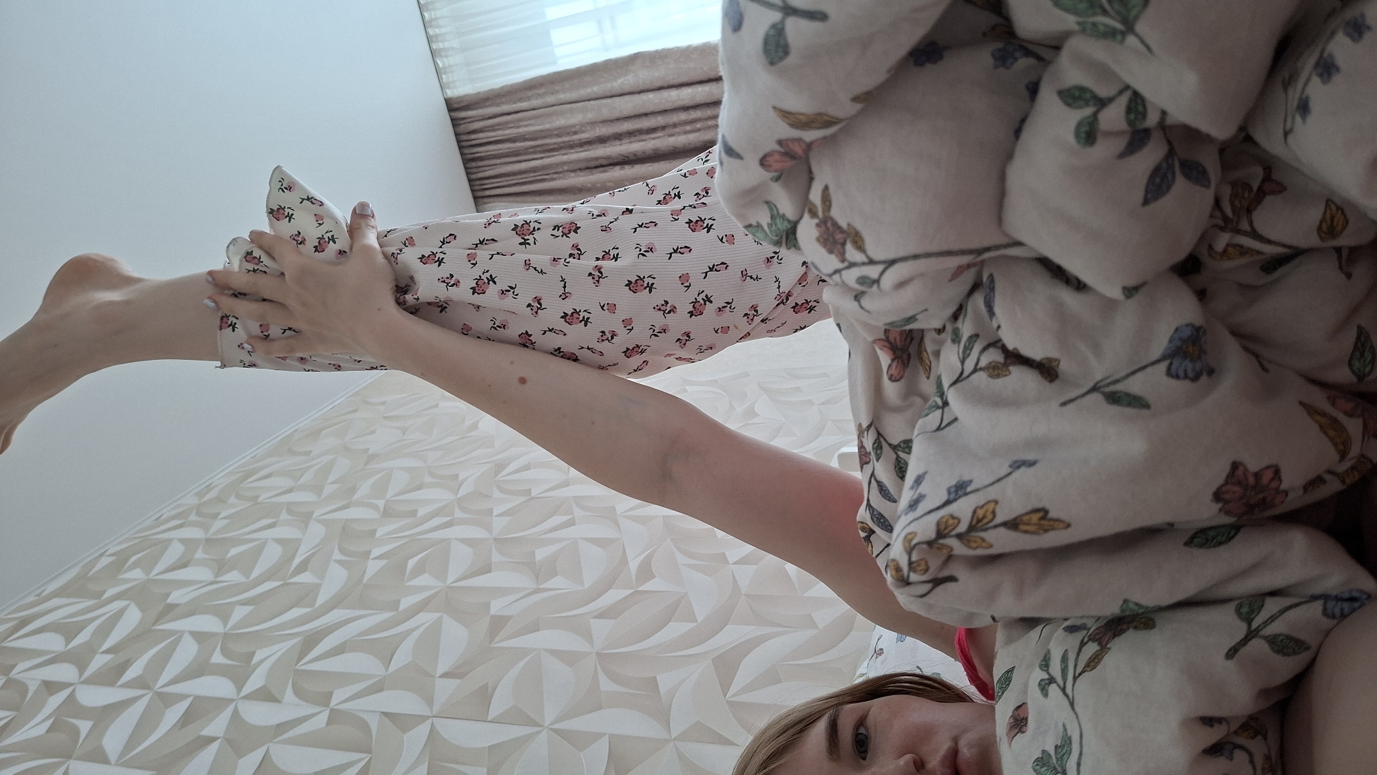 A woman lies in bed in a room in Chaikovsky, Russia. She is partially covered by a floral-patterned duvet and is wearing floral-patterned pajamas. She is holding her leg up in the air with both hands. The room has textured white walls and a window with a curtain.
