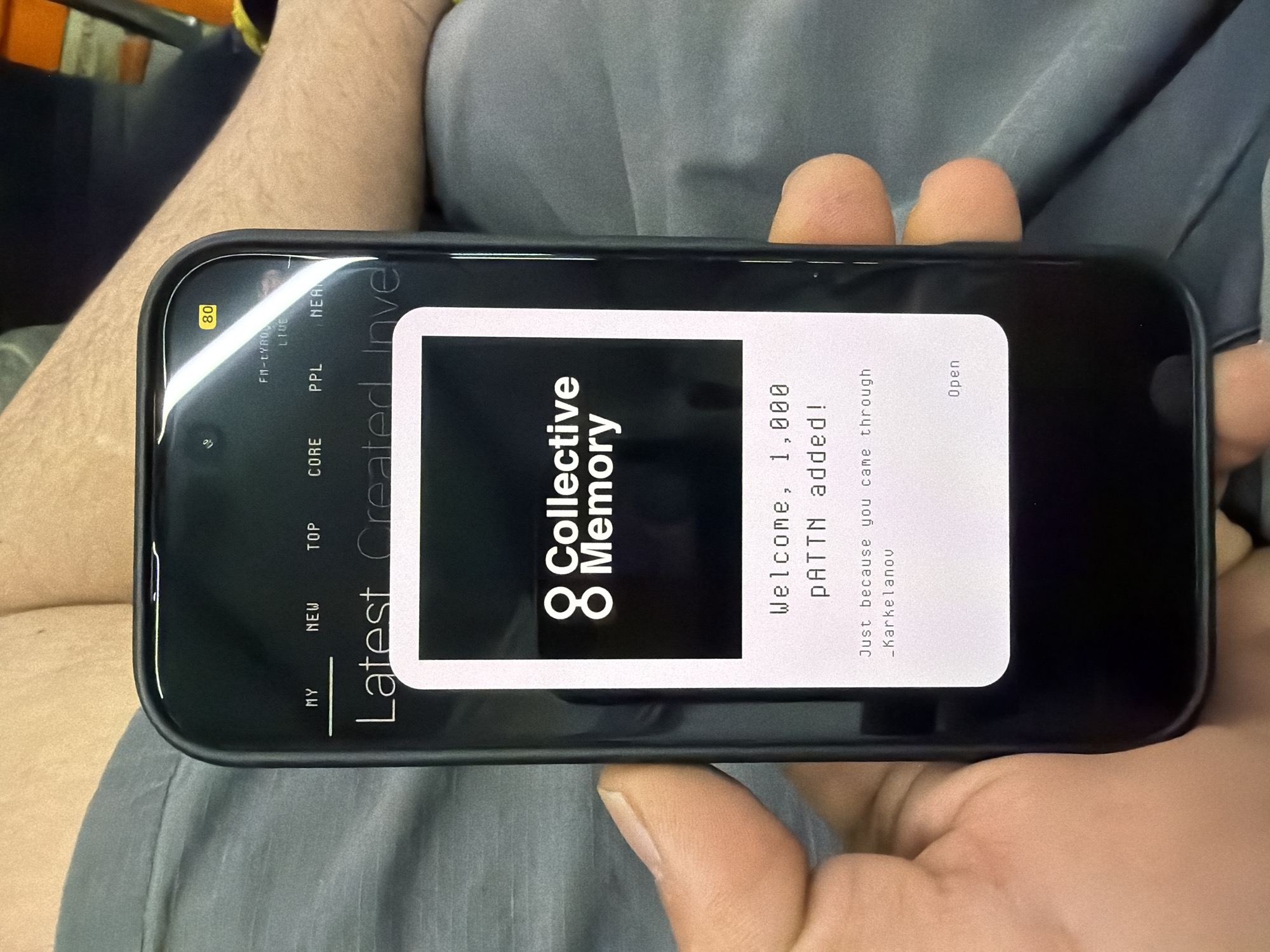The image depicts a person holding a smartphone. The phone's screen displays the app "Collective Memory" with a welcome message indicating "1,000 PATTN added." The screen also includes navigation tabs: "MY," "NEW," "TOP," "CORE," "PPL," "LIVE" and "Latest Created Investments". The upper left corner of the phone shows "80" in a yellow box. The person holding the phone is wearing grey pants. There is no information about the location in Burgas, Bulgaria.