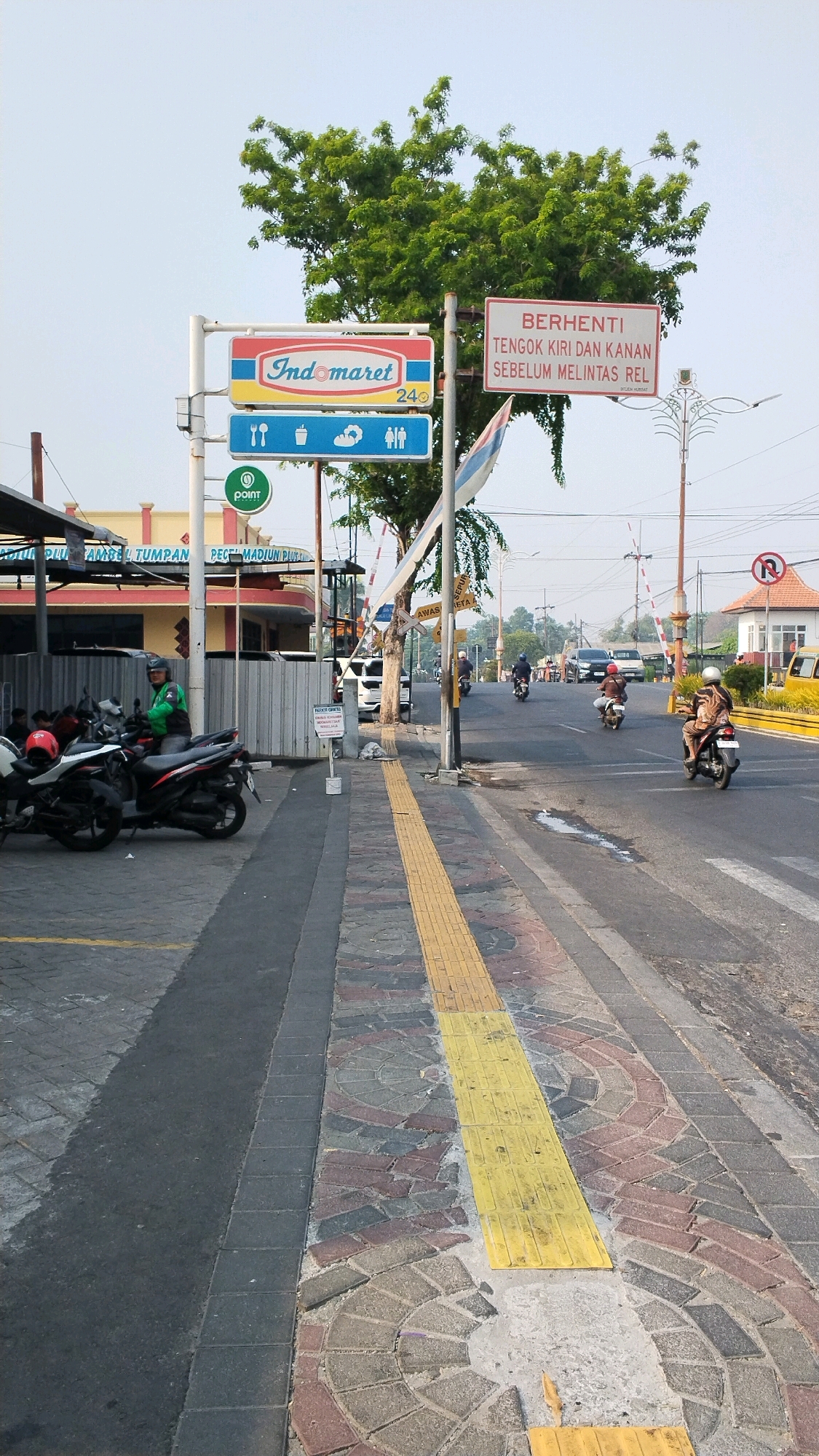 The image captures a street scene in Mojokerto, Indonesia, near a railroad crossing. A prominent Indomaret convenience store sign is visible, along with a sign indicating "BERHENTI TENGOK KIRI DAN KANAN SEBELUM MELINTAS REL" (Stop, look left and right before crossing the tracks). A train crossing gate is lowered, with a red and white barrier visible. Several motorcycles are parked on the sidewalk, some with riders. One rider, wearing a green jacket and helmet, is seated on a motorcycle.
The sidewalk features a textured yellow tactile paving strip, indicating a path for visually impaired pedestrians. The street is paved with asphalt, and there are some puddles of water on the road surface. Several vehicles, including motorcycles and cars, are present on the road, some in motion. In the background, trees line the street, and there are buildings on the side, including a structure with a red roof. The sky is clear and bright, suggesting daytime. The overall scene depicts a busy urban environment with typical Indonesian street elements.