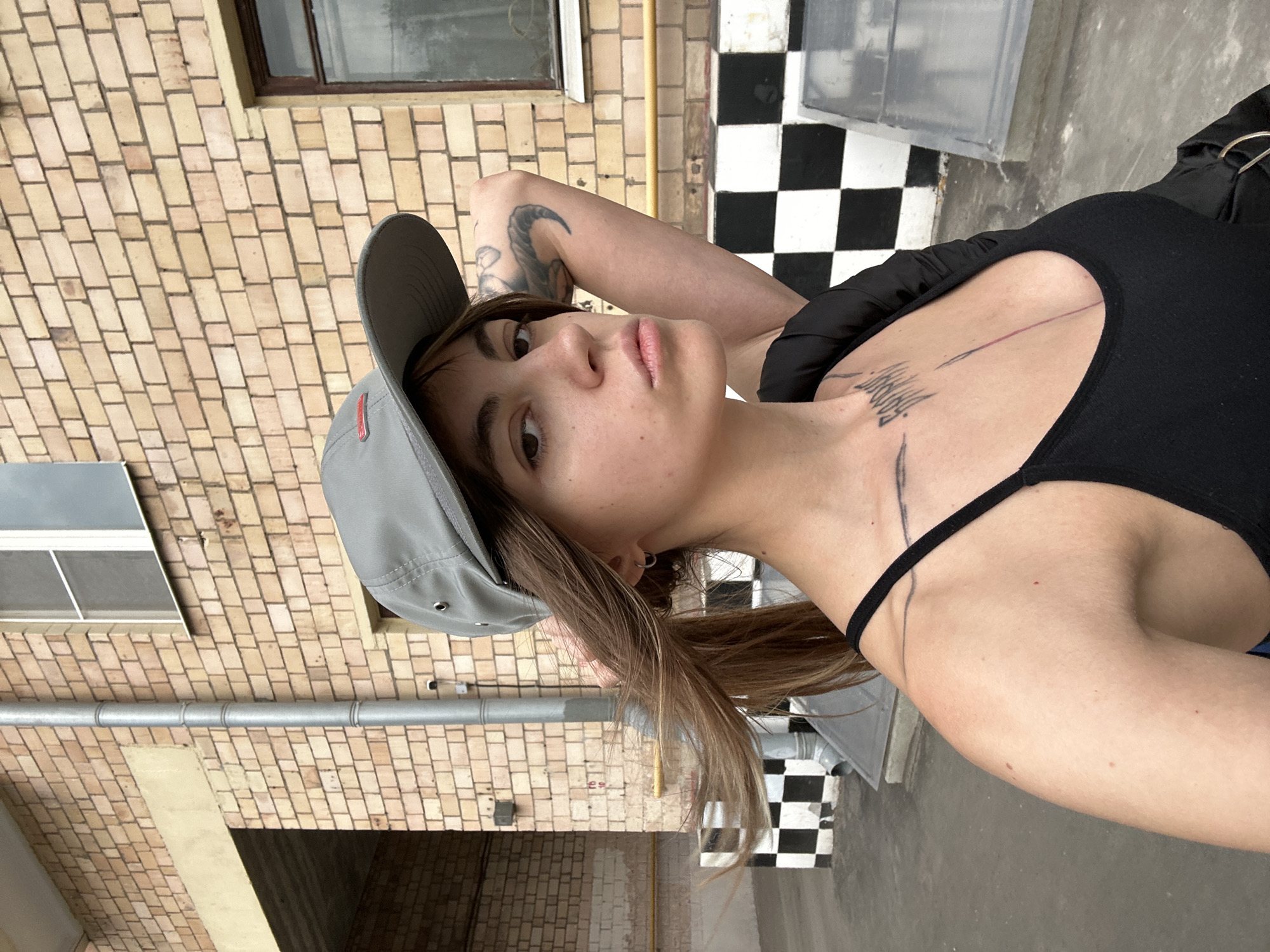 A young woman wearing a grey baseball cap, a black tank top, and a black jacket stands outside in front of a brick building with a checkered pattern on the wall. The woman has long brown hair, a tattoo on her arm, and a tattoo on her chest. She is holding her hair back with her hand. The image appears to have been taken in Moscow, Russia, based on the presence of a Moscow Metro station sign in the background.