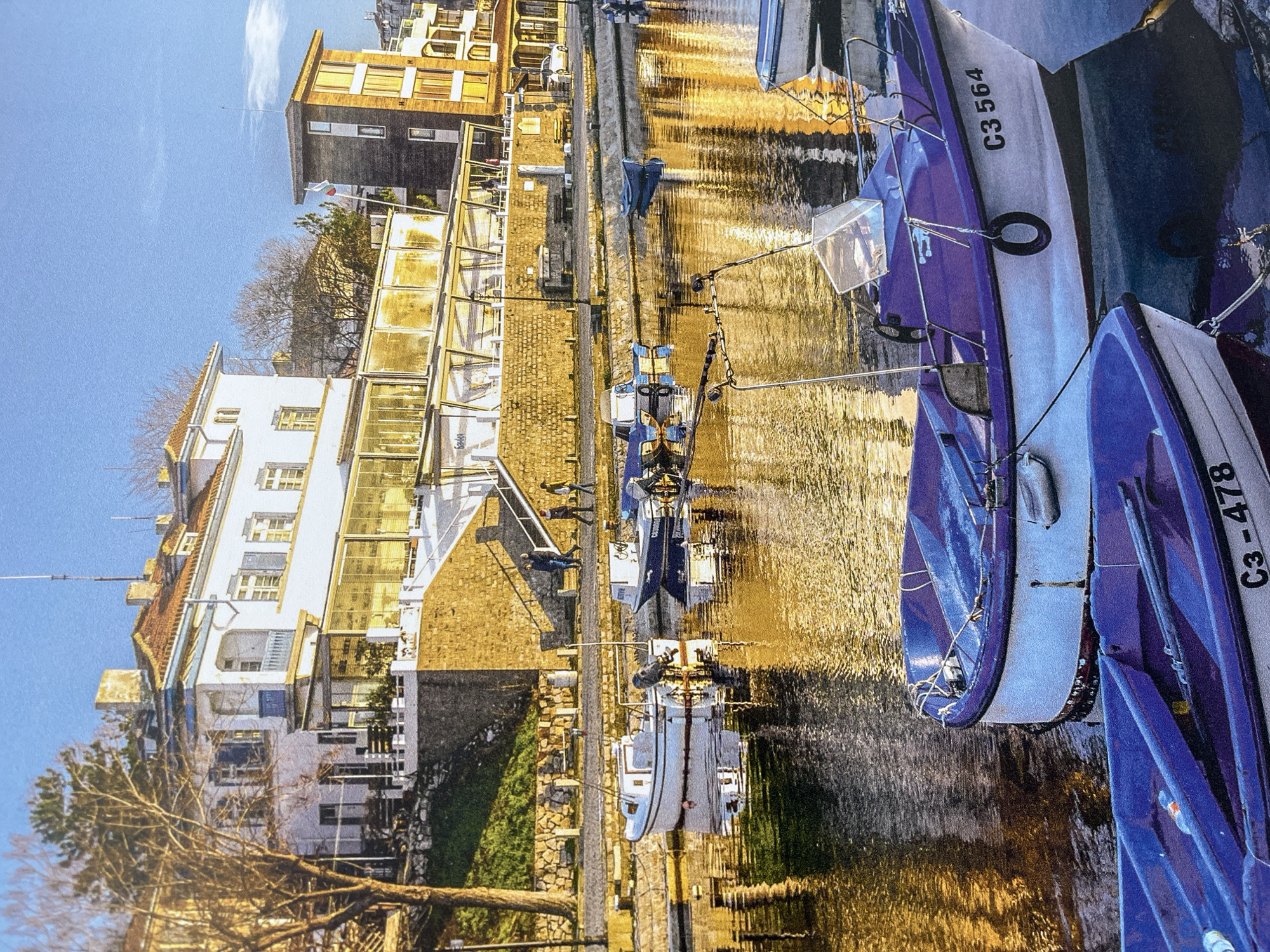 The image depicts boats docked in a canal in Burgas, Bulgaria. The boats, painted white and blue, are registered with codes "C3-478" and "C3-564." The water reflects the sunlight and the buildings on the canal's edge. Along the canal, there's a stone walkway with people walking on it. Buildings on the other side of the canal display varying architectural styles. A tree is visible in the foreground.