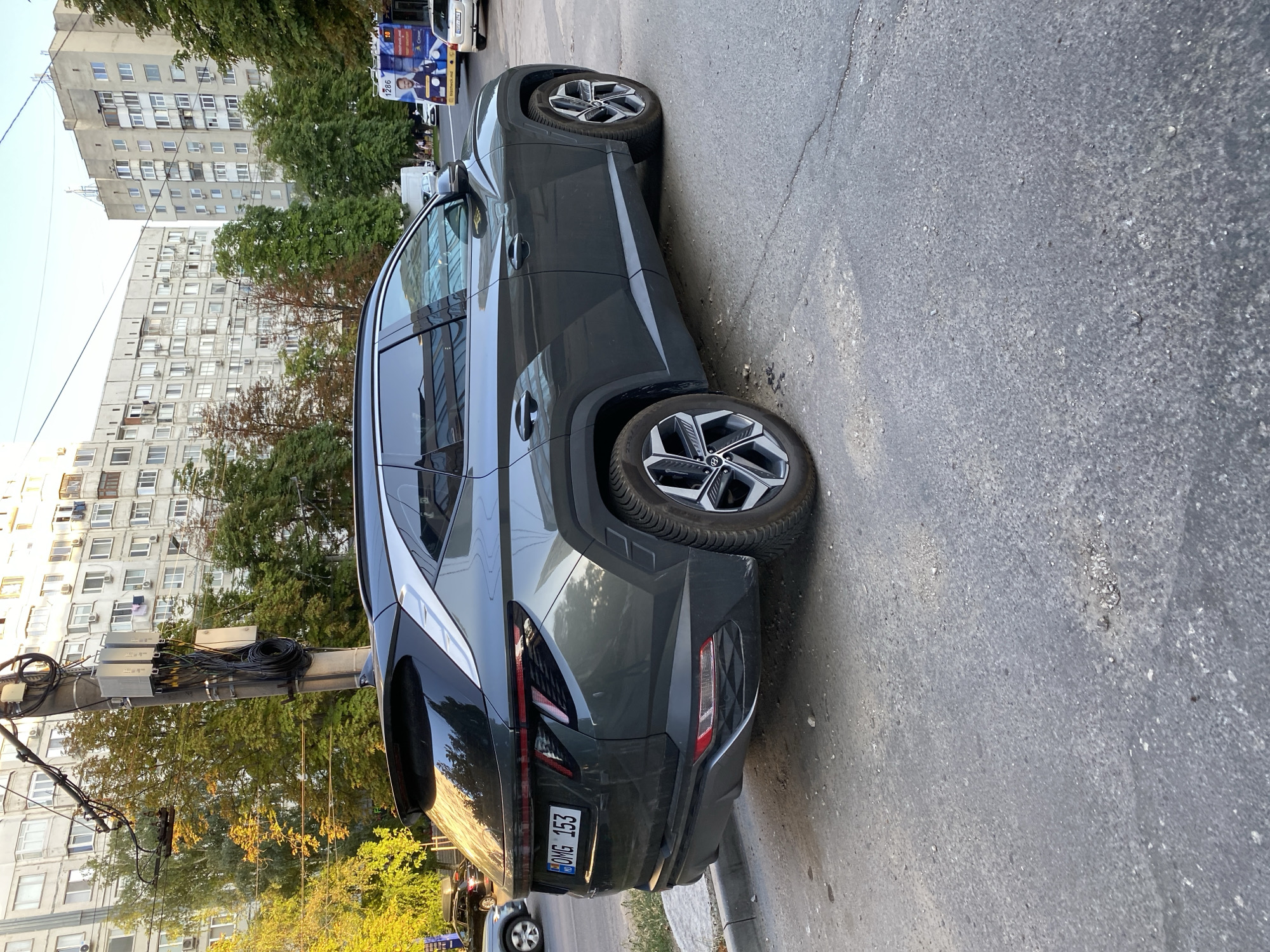 A dark gray SUV, possibly a Hyundai Tucson, is parked on a street in Chișinău, Moldova. The vehicle's license plate reads "OMG 153". It is daytime, and the sun is casting shadows. In the background, several apartment buildings are visible, typical of Soviet-era architecture, with visible air conditioning units on the facades. Trees with some autumn foliage are also present. A utility pole with numerous cables and communication equipment is situated near the car. Further down the street, a bus with advertisements and people can be seen. The street surface appears to be asphalt with some cracks and debris.