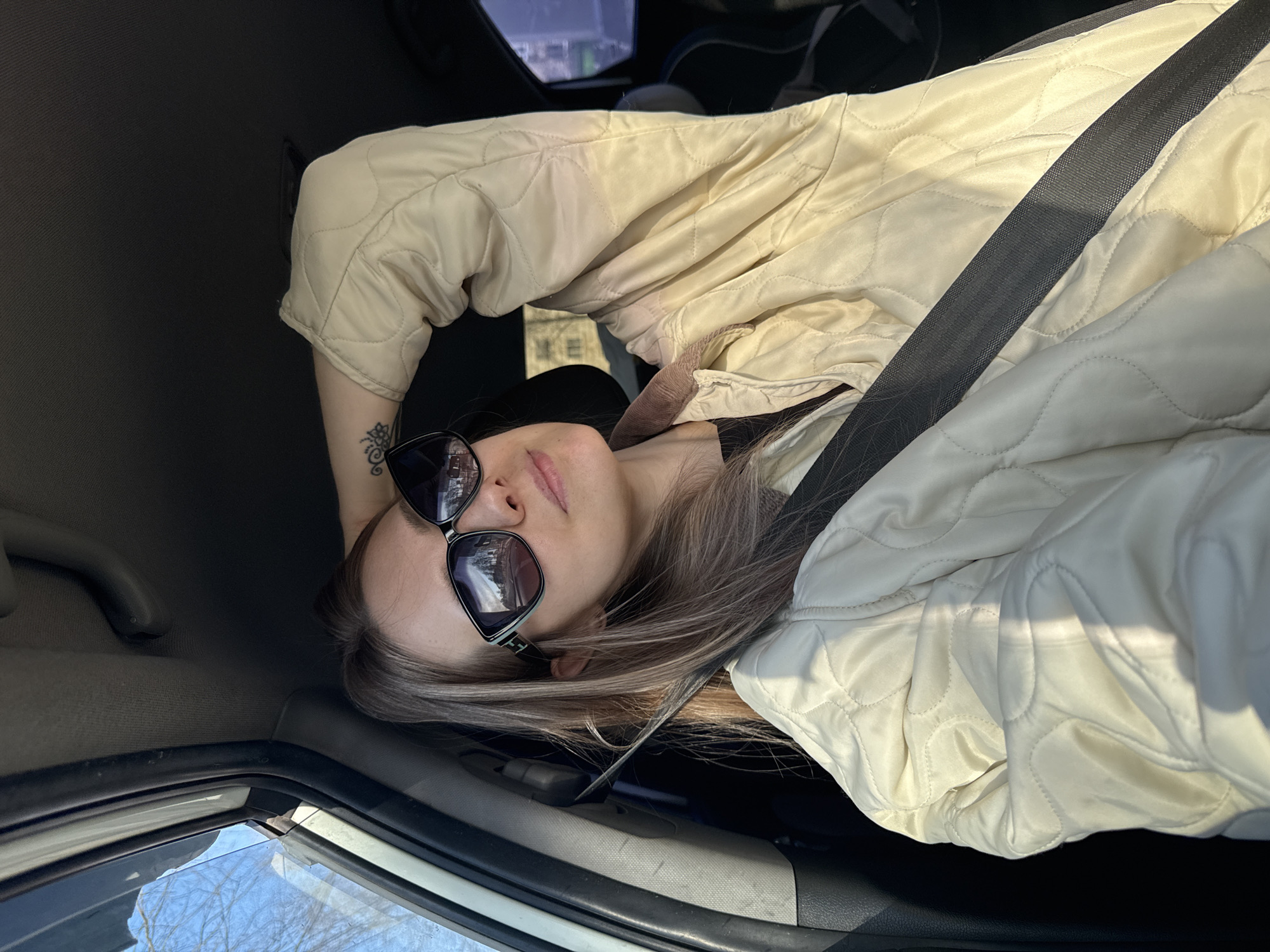 A woman wearing sunglasses and a light-colored quilted jacket with a seatbelt across her body is reclined inside a car. The scene is lit by daylight. Outside the car window, bare trees are visible. The woman has a tattoo on her left wrist. The reflection in her sunglasses shows buildings and other details of an urban setting. While the location cannot be explicitly verified as Chaikovsky, Russia from the image alone, the details suggest an urban environment that could plausibly be in that city.