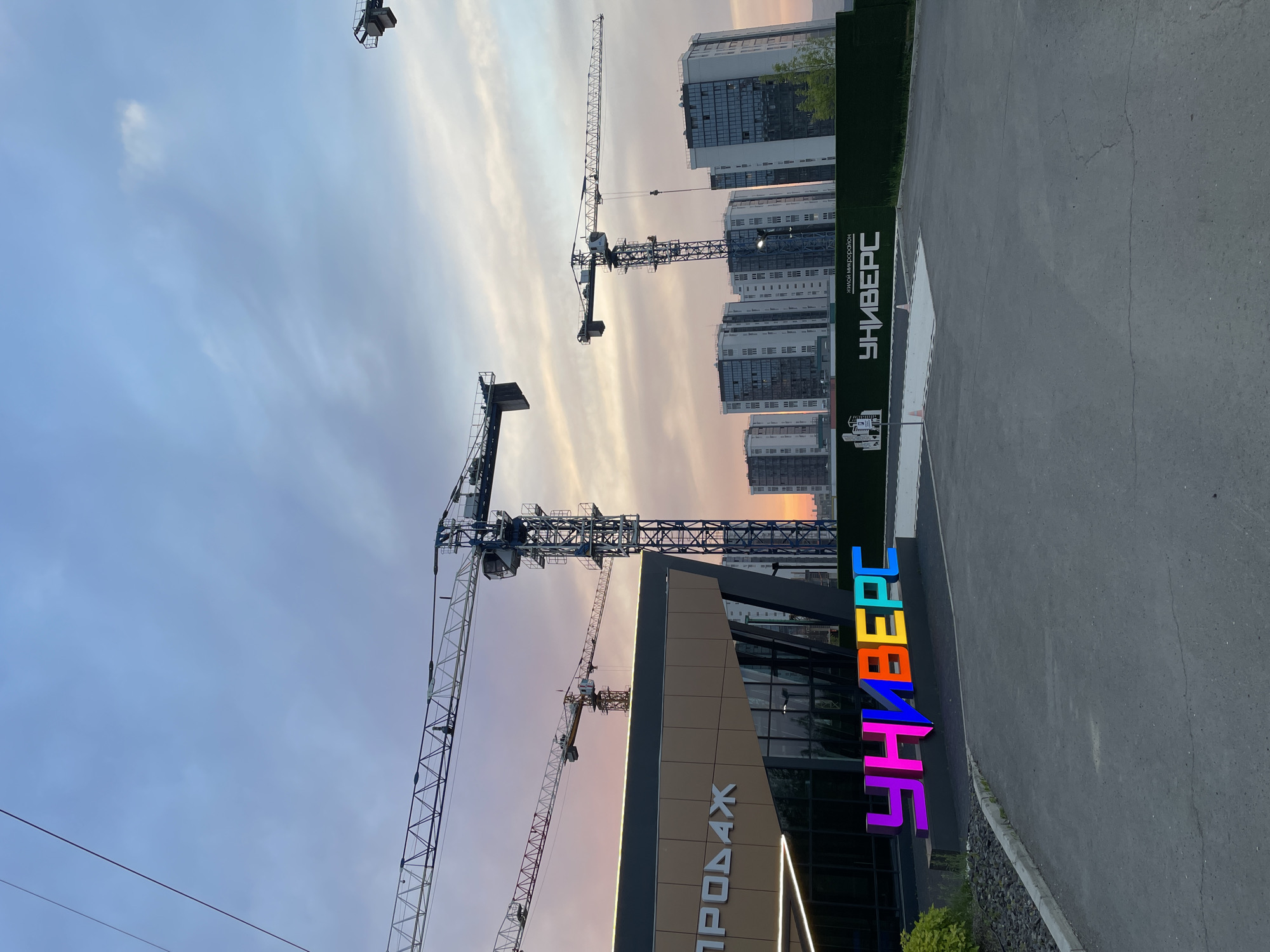 The image shows a construction site in Krasnoyarsk, Russia. Several construction cranes are visible against a twilight sky. In the foreground, a building with "ПРОДАЖ" (Sales) written on it and a large, colorful "УНИВЕРС" sign are present. In the background, apartment buildings under construction and a green fence with "ЖИЛОЙ МИКРОРАЙОН УНИВЕРС" (Residential District UNIVERS) are visible. There are no people or animals in the image. The scene appears to be a developing residential area.