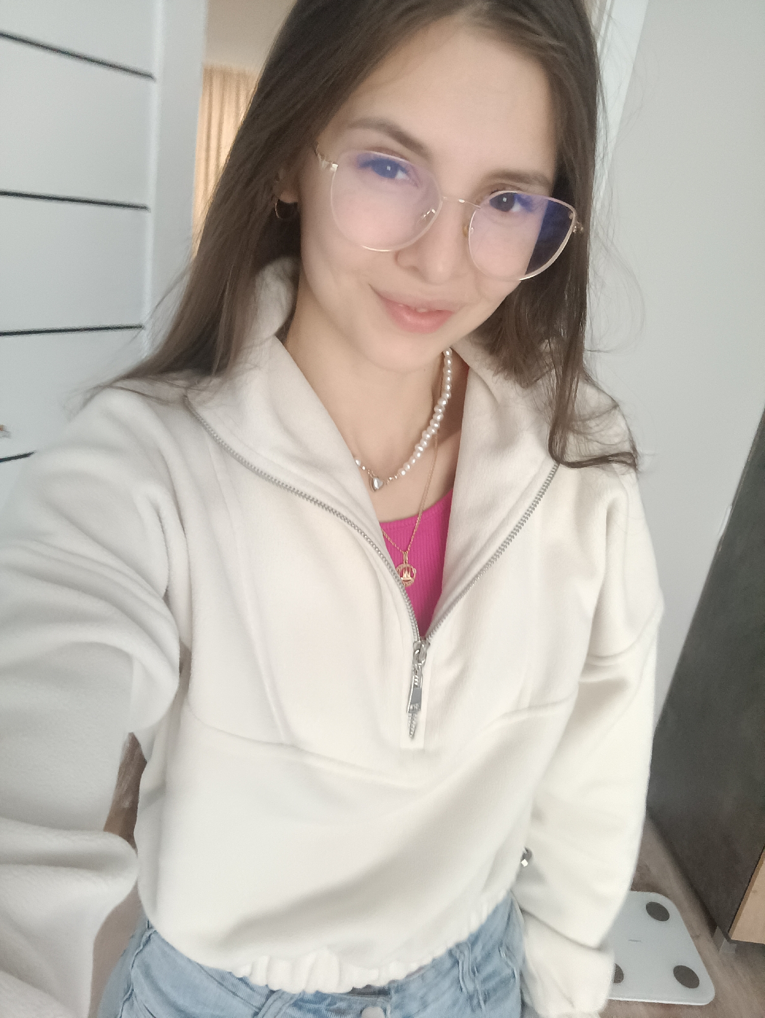 A young woman with long brown hair and glasses is taking a selfie indoors. She is wearing a white, quarter-zip fleece jacket over a pink top, a pearl necklace, and jeans. A second necklace with a pendant is also visible. Behind her is a white door with horizontal lines, and a partially visible beige curtain. To her right, a dark gray object is present on the floor, with a white scale visible as well. The location is Unknown, Unknown.