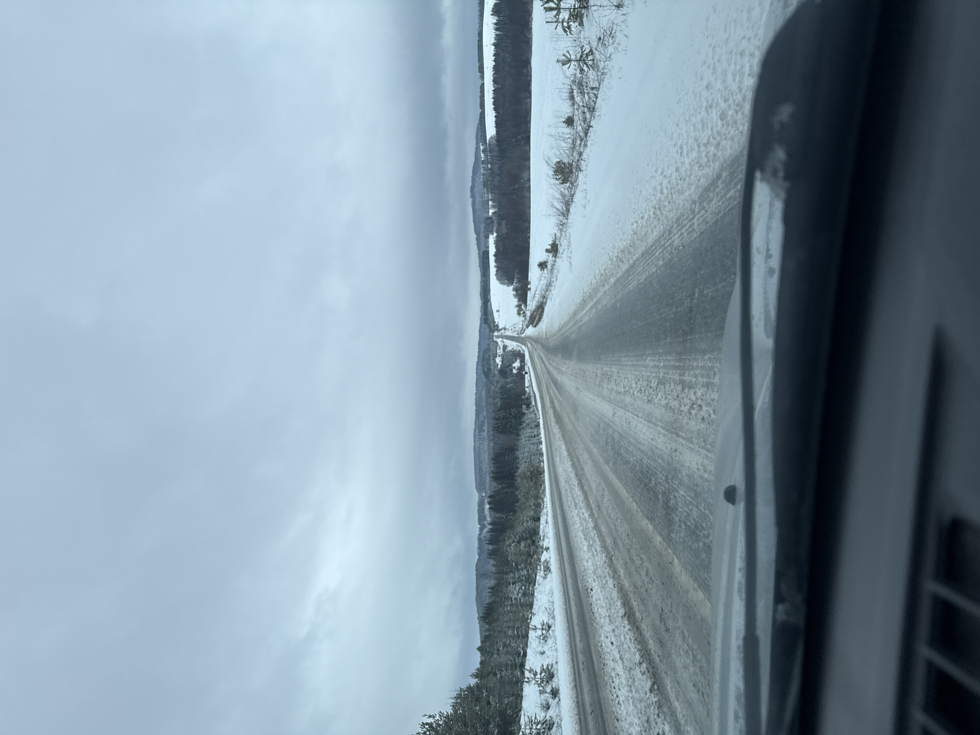 A long, straight, snow-covered road stretches into the distance, flanked by snow-covered fields and wooded areas. The sky is overcast. The photo appears to be taken from inside a vehicle moving along the road. The location is possibly near or around Samolet, Russia, based on the prompt. No people are visible. The primary objects are the road, snow, fields, trees, and the interior of a vehicle. The road appears to be traveled, as tire tracks are visible.