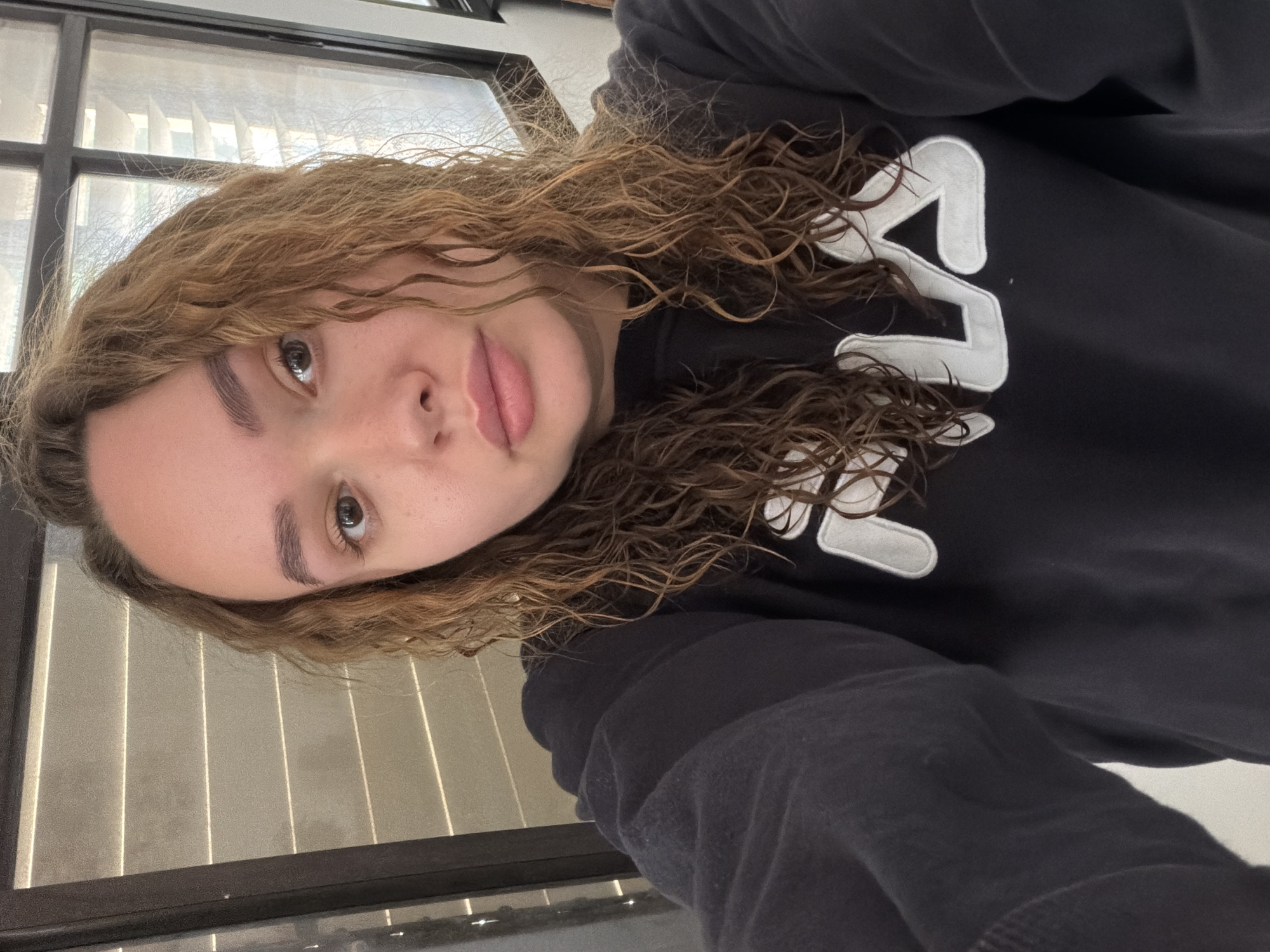 A young woman with light brown, curly hair is wearing a black Fila sweatshirt and posing for a selfie. She has fair skin, brown eyes, and full lips. The background shows a window with blinds. The location cannot be determined from the image.