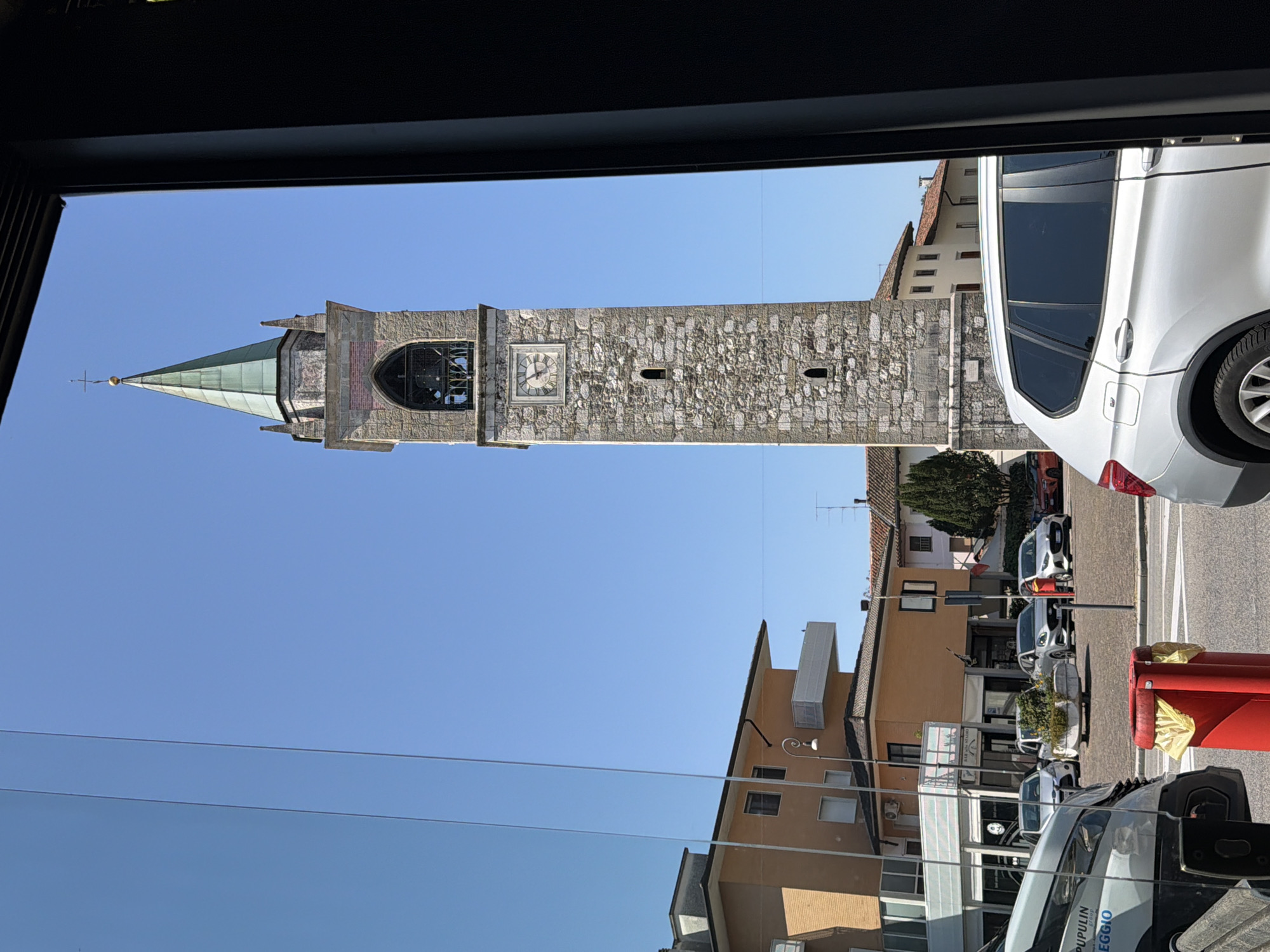 The image shows a street scene in the city of Udine, Italy, on a clear, sunny day. In the background, a prominent stone bell tower with a green steeple dominates the skyline. The tower has a clock face, arched openings, and is constructed of rough-hewn stones. To the left of the tower, several buildings are visible, including a light brown, multi-story building with balconies and windows, and a lower, darker building that appears to be a shop with signage. In the foreground and mid-ground, cars are parked and driving on a paved road. A silver SUV is parked on the right side of the image. A white van or truck is on the left, with some text visible on its side reading "PUPULIN GROUP LEGGI." A bright red trash bin with yellow bags is positioned on the road. The overall impression is of a typical urban street with historical architecture.