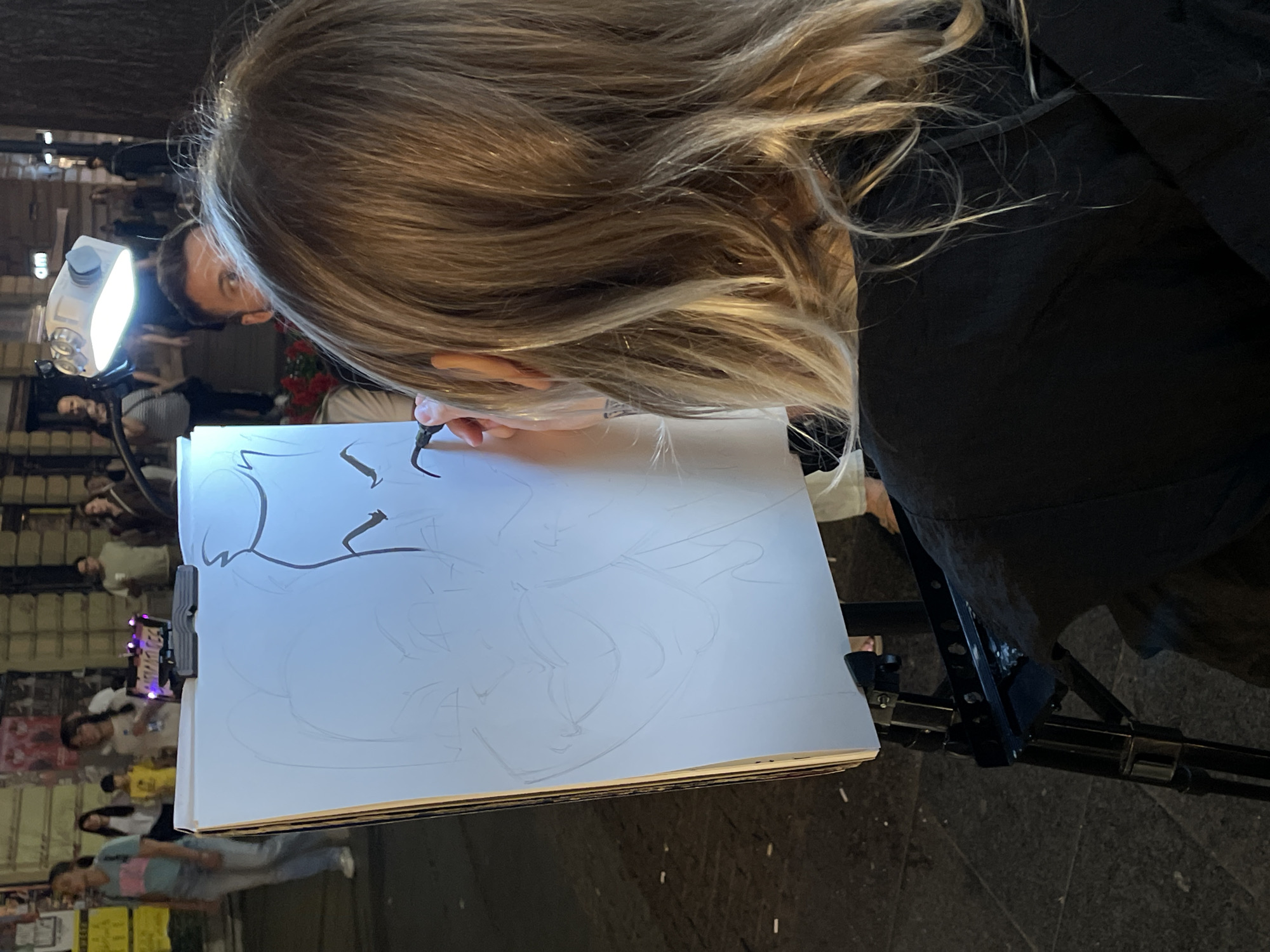 A woman with blonde hair is bent over a drawing on a white pad attached to a black metal easel. She holds a pen in her right hand, sketching on the paper, illuminated by a small white lamp attached to the easel. The drawing appears to be a stylized, cartoonish portrait. To the left, out of focus, a group of people are standing or walking along a street. The ground appears to be paved with gray stone. Based on other images of this subject, the location is likely in Belgrade, Serbia.