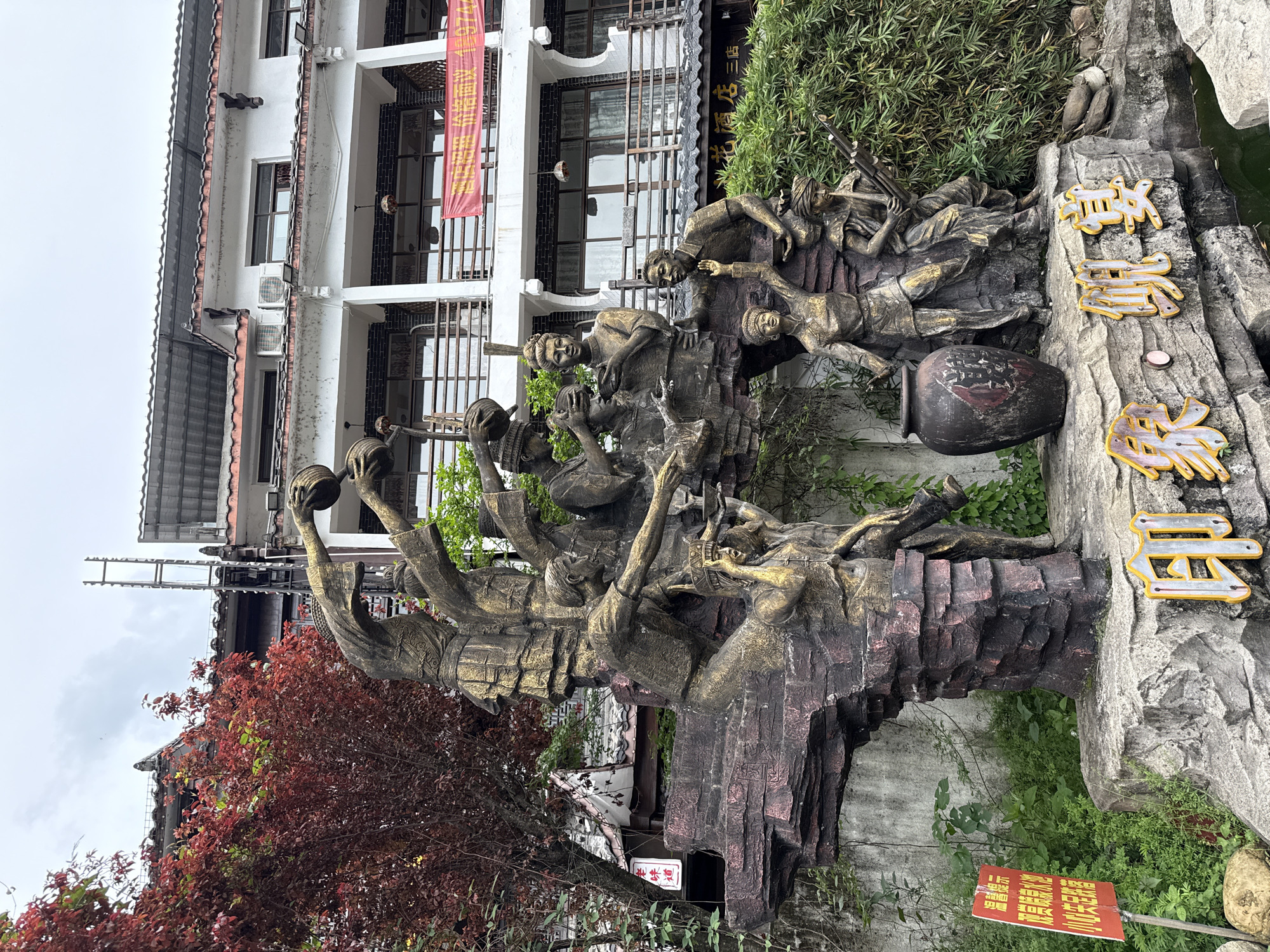 The image captures a large, intricate outdoor sculpture set against a building and natural landscaping, likely in Wulingyuan, China. The sculpture, rendered in a dark, weathered material with gold accents, depicts numerous human figures engaged in a dynamic, collective activity. The figures appear to be celebrating, working, or performing a ritual, with some holding bowls or baskets aloft, others gesturing, and one lying down holding a long object, possibly a musical instrument or tool. A large, dark pot or jar is integrated into the right side of the sculpture's rocky base.
Behind the sculpture, a multi-story building with white walls, dark window frames, and traditional roof tiles is visible. A red banner hangs from the building, displaying Chinese characters that read "湘西味道" (Xiangxi Taste/Flavor), suggesting a restaurant or food-related business. Lush green bamboo-like plants and other shrubs grow behind and around the sculpture, and a tree with vibrant red foliage stands to the left. A small water feature or pond can be seen on the far right.
Prominently displayed on the rocky outcrop to the right of the sculpture are three stylized Chinese characters with gold outlines and pearlescent infill, reading "溪布街" (Xibu Jie), indicating the name of the street or area. A small red sign on the ground in front of the sculpture warns "禁止攀爬" (No Climbing). The overcast sky suggests daytime. The overall scene depicts a cultural or historical commemoration embedded within an urban, yet naturally integrated, environment.
