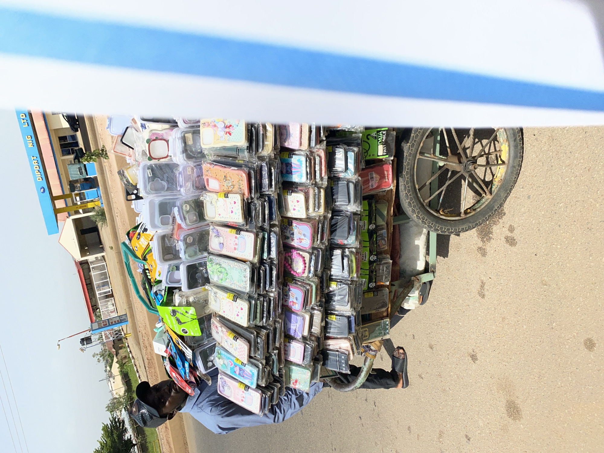 An overhead shot captures a street vendor, a Black man wearing a blue shirt and a black cap, standing next to a cart laden with phone cases. The cart is filled with rows of phone cases, organized by color and design. The vendor's feet are visible, clad in black flip-flops, on the asphalt street. In the background, a gas station with the "DIPDAT NIG LTD" sign is visible, along with buildings and trees, suggesting an urban or suburban setting. The sky is bright and clear, indicating a sunny day. The scene is set in Bukuru, Nigeria.