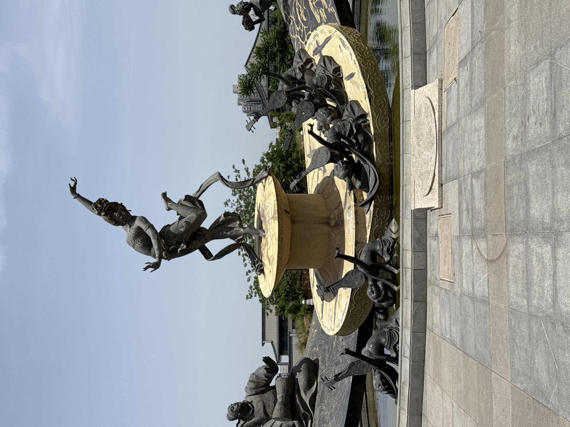 The image captures a detailed, bronze sculpture located in Lintong District, China. The sculpture features a central figure of a dancer in mid-leap with one arm extended skyward. The dancer has long hair, jewelry, and a garment around the torso. The sculpture includes several layers or levels, with figures holding musical instruments around a golden platform. A pool of water is situated nearby, and a paved walkway extends into the foreground. Behind the sculpture, trees and a multi-story building can be seen in the background. The sky is a light blue color.