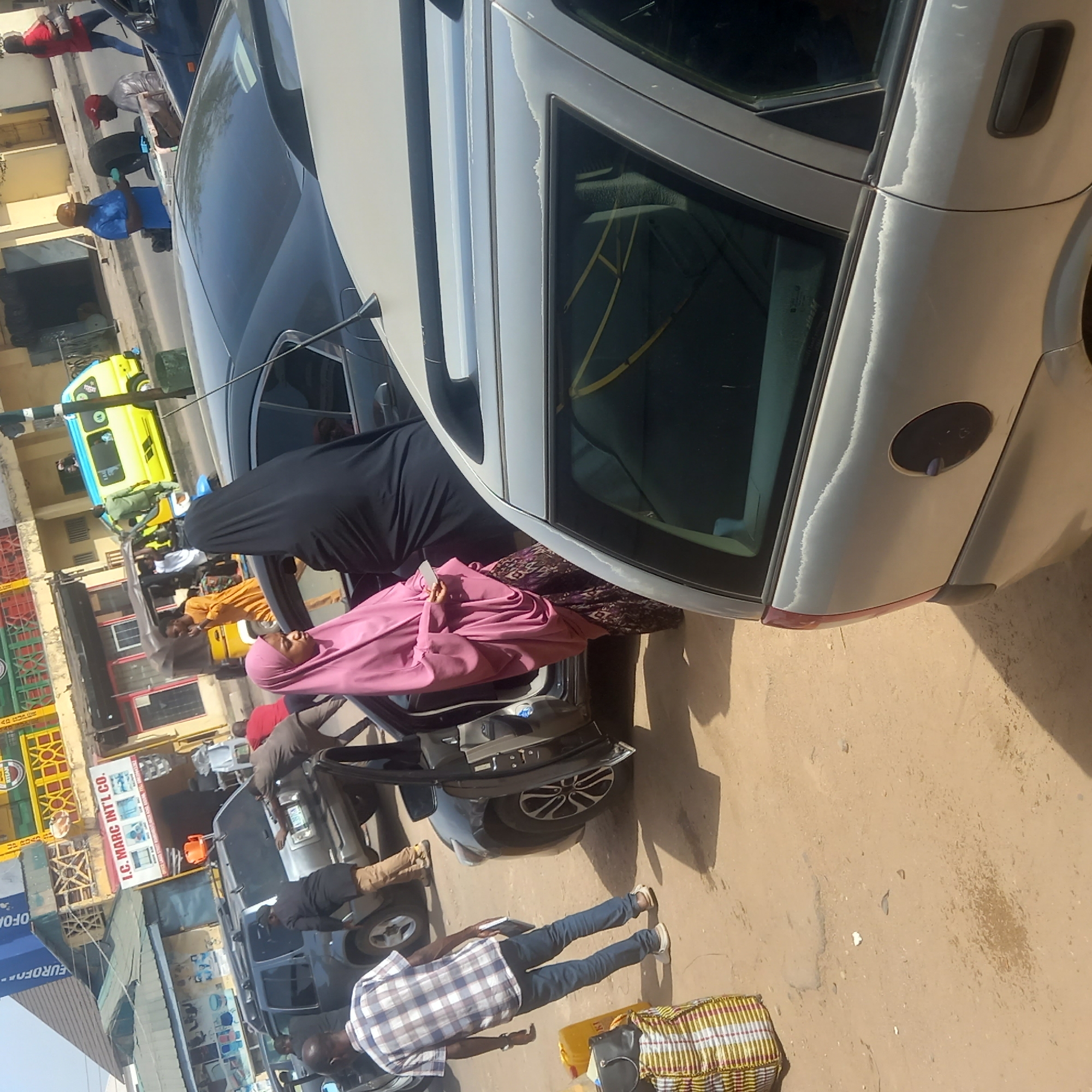 This image captures a bustling street scene in Dadi Riba, Nigeria, under bright daylight. The primary focus is on a busy roadside area with several people and vehicles.
In the foreground, a silver car, likely a hatchback or wagon, is prominently featured. The car's paintwork shows significant wear and peeling, particularly along the side windows and roof edges. Next to this vehicle, a woman dressed in a black abaya or long dark garment stands near an open car door. Another woman, wearing a pink hijab and corresponding attire, is either entering or exiting the car.
Adjacent to the silver car, a dark-colored car, possibly a sedan or SUV, is parked with its rear passenger door open. A man in a plaid shirt and blue jeans stands next to this vehicle, with a distinct striped, woven bag and a yellow bag placed on the dusty ground nearby. Further back, another man in a blue shirt and a cap is visible near a tire. Several other individuals are scattered throughout the scene, some appearing to interact with vehicles or simply standing.
The midground features more vehicles, including what looks like a grey truck or SUV. In the background, typical commercial buildings line the street. A bright yellow and blue minibus, commonly used for public transport in Nigeria, is visible further down the road. Visible text on a white sign with blue letters reads "I.C. MARC INT'L CO." Another blue sign partially shows "EUROFO...". The ground is unpaved and appears sandy or dusty. The strong sunlight casts clear shadows, indicating a sunny day. The overall impression is one of active daily life in a vibrant urban or semi-urban setting.