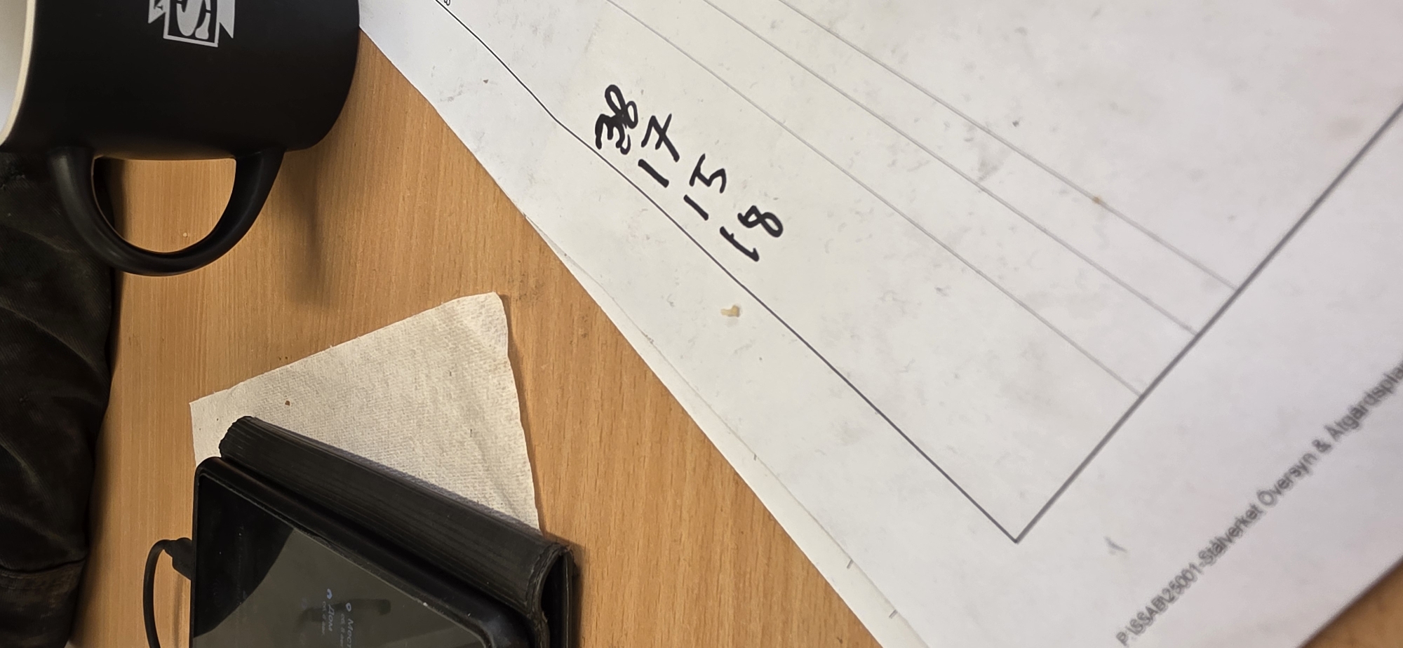 The image shows a close-up view of a wooden desk. On the desk, there is a black tablet in a black case, a white paper napkin, and a black coffee mug with a white logo. A document with handwritten numbers is also visible. The numbers written on the document are 38, 17, 15, and 18, written vertically. The bottom left corner of the document shows the text "P.ISSAB25001-Ståverket Översyn & Åtgärdsplan". This document information indicates the photo was taken at a location in Luleå, Sweden, specifically within the "Ståverket" (steelworks) facility, and relates to an inspection and action plan. The overall scene suggests a work environment, possibly during a break, given the presence of the coffee mug and tablet.