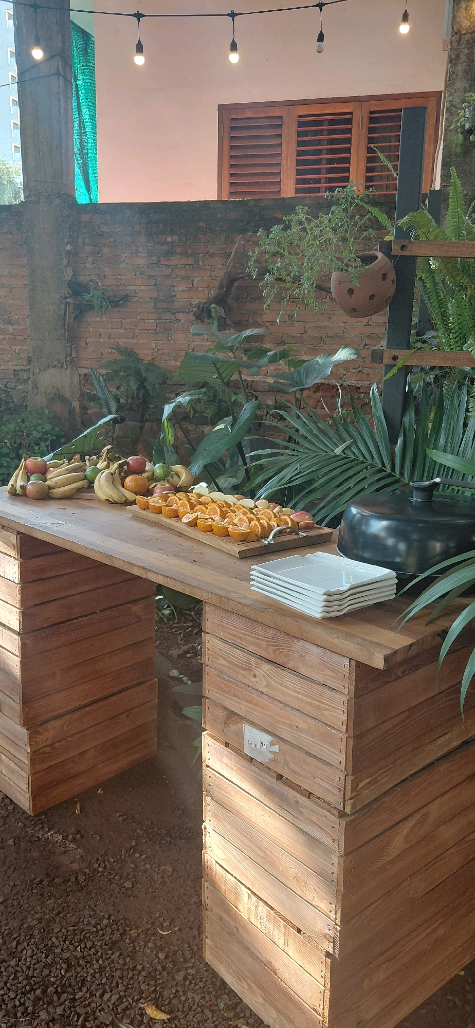 A close-up, eye-level, outdoor shot captures a wooden table laden with a variety of fruits, set against a backdrop of a brick wall and lush greenery. The table, constructed from stacked wooden crates, appears to be located in a garden or patio area in Encarnacion, Paraguay. On the table, a colorful assortment of fruits includes bananas, apples, oranges, and possibly some grapes or berries. A large, dark pot with a lid sits on the right side of the table, possibly a serving dish or cooking vessel. To the right of the fruits, a stack of white square plates is neatly arranged, suggesting a meal or gathering. The environment is verdant, with large-leafed plants and ferns surrounding the table, hinting at a tropical or subtropical setting. The brick wall suggests an older structure, possibly a part of a residential property or a restaurant's outdoor seating area. String lights are visible overhead, indicating this space might be used for evening events. The ground is covered with small, dark gravel. The lighting suggests it is daytime, with sunlight casting shadows on the wooden crates and highlighting the textures of the fruits and greenery. No people are visible in the frame.