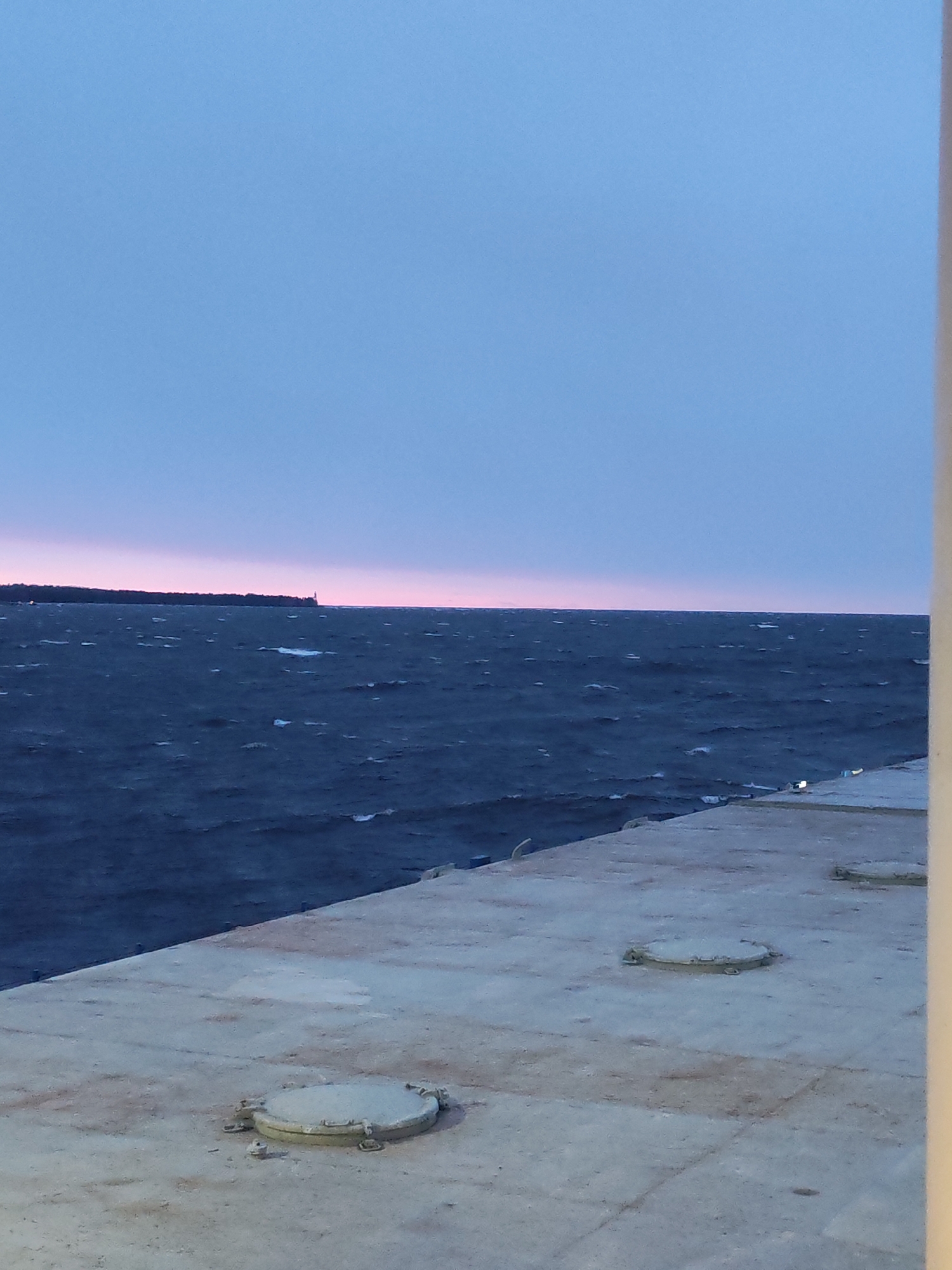 The image captures a wide shot of a twilight scene over a large body of water, viewed from a concrete structure. The sky, occupying the upper half of the frame, transitions from a pale blue at the top to a band of soft pink and orange hues along the horizon, indicative of either sunrise or sunset. The body of water, likely a sea or a large lake, appears dark blue-grey and is visibly choppy with numerous whitecaps, suggesting windy conditions.
Along the distant horizon, a dark, tree-lined landmass is discernible, with a slender, white structure—likely a lighthouse—standing prominently. The lower part of the image features a light-colored, textured concrete surface, possibly a pier, dock, or ship deck, extending from the bottom left corner towards the right. Two circular, hatch-like metal covers are embedded in this concrete surface. A narrow, off-white vertical element is visible along the far right edge of the frame.
There are no people or explicit activities depicted. The scene conveys a sense of serene solitude, dominated by the expanse of water and sky under the low light conditions. The weather appears breezy, evidenced by the rough water. The location is likely a coastal area or a large inland body of water, such as near Shcheleiki, Russia. No visible text is present in the image.