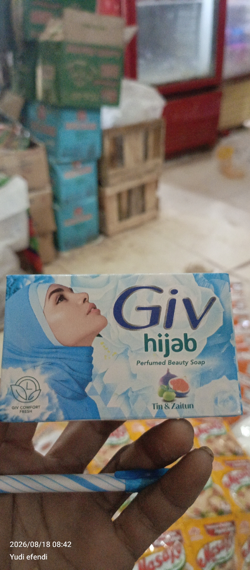 In Rawajitu Selatan, Indonesia, a close-up image captures a package of Giv hijab perfumed beauty soap. The soap packaging features a portrait of a woman wearing a blue hijab and a floral design. Below the brand name, "Giv hijab," it is described as "Perfumed Beauty Soap." The packaging also displays an image of figs and olives, with the text "Tin & Zaitun" beneath it. In the bottom left corner, a circular logo with leaves and a "L" inside is visible, with the text "GIV COMFORT FRESH." The image also includes a timestamp "2026/08/18 08:42" and the name "Yudi efendi." The background is blurred, showing stacked boxes and shelves, suggesting a retail or storage environment. The soap package is held by a person's hand, partially visible at the bottom of the frame.