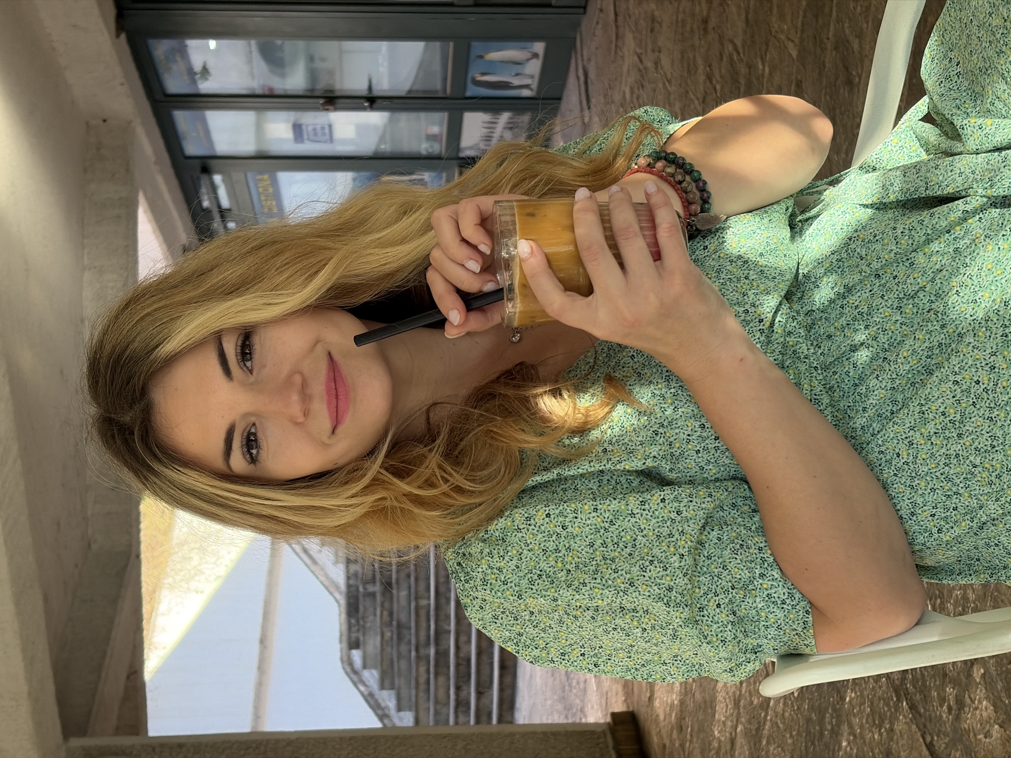 A young woman sits at an outdoor table, holding a beverage with a straw. She is wearing a green floral dress and bracelets. The background features a store window with reflections and stairs leading to an upper level. The scene is likely located in an outdoor cafe or seating area in Budva, Montenegro, given the style of architecture and materials visible in the image.