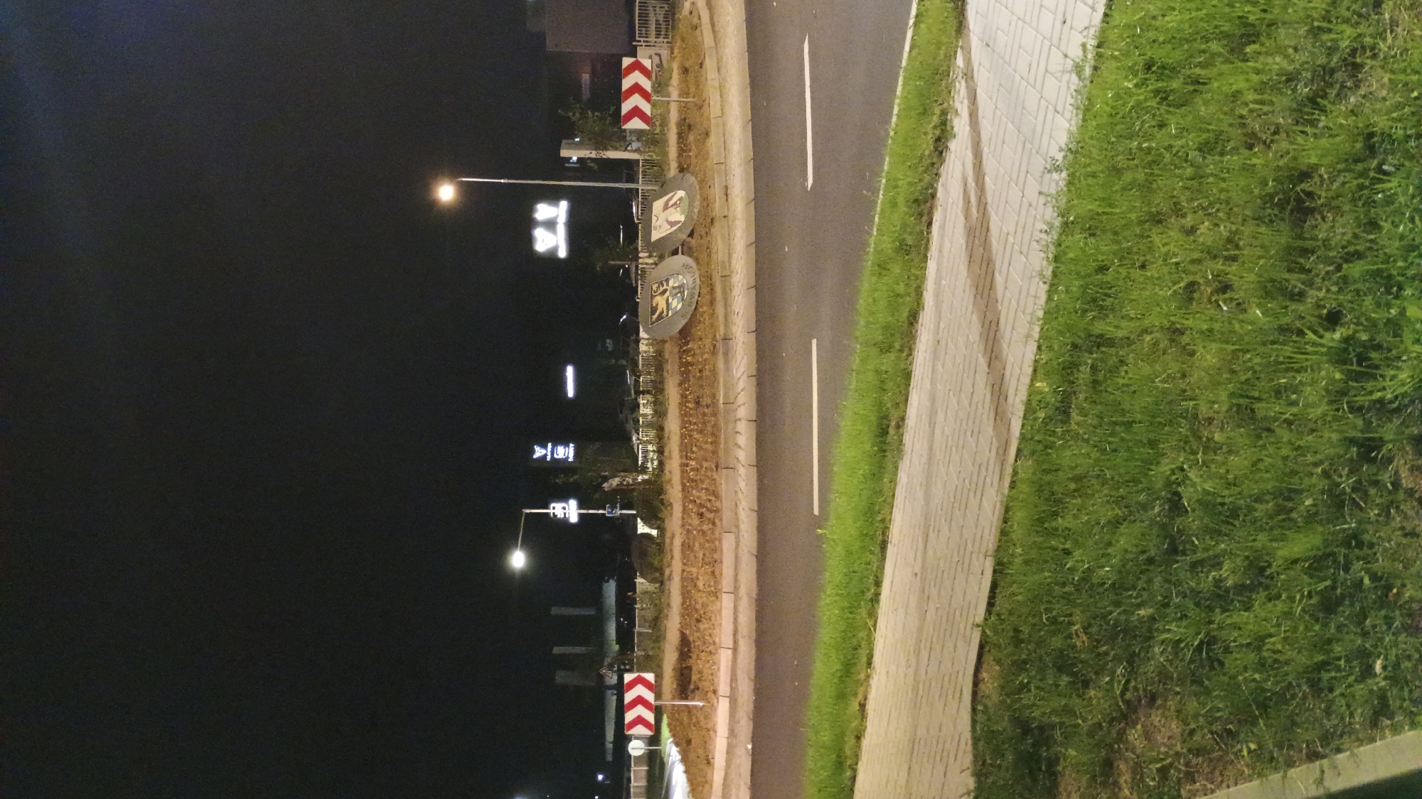 A nighttime photograph captures a roundabout in Sokolov, Czechia. A street lamp illuminates the scene, casting light on a circular island at the center of the roundabout. Two large, circular emblems, possibly coat of arms, are placed on the island, facing outwards. Red and white chevron signs are visible on the approaches to the roundabout, indicating traffic direction. The roadway is asphalt, with white lane markings. A paved pedestrian walkway runs alongside the road, bordered by a strip of green grass. In the background, various buildings and illuminated signs are faintly visible against the dark night sky. No people or vehicles are present in the immediate vicinity of the roundabout.