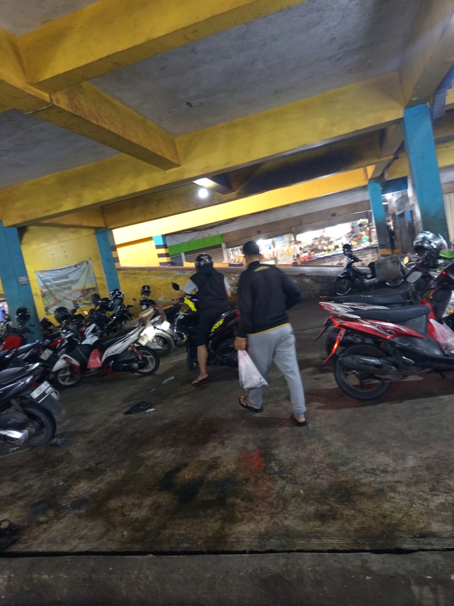 This image depicts a covered outdoor parking area, likely at a market or public facility in Karawang, Indonesia. The scene is characterized by a concrete floor, a yellow-painted ceiling with exposed beams, and blue and yellow support columns. Numerous parked motorcycles, predominantly scooters, are visible, suggesting a busy location. In the foreground, two men are walking. One man, wearing a black vest over a grey long-sleeved shirt and grey pants, carries a white plastic bag. The other man, wearing a black jacket with yellow accents and grey pants, is also walking away from the viewer. The lighting suggests it is either early morning or late evening, or the area is under artificial light. The overall impression is one of a functional, well-used public space.